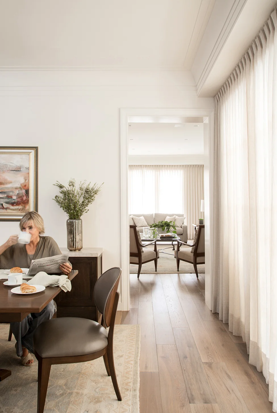 Woman drinking coffee at a wood dining table with a view of the living room