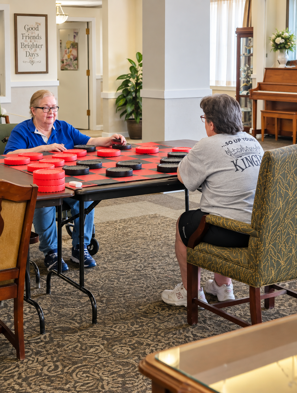 Two adults are playing checkers at a table in a living room or common area. The woman on the left, with blonde hair and glasses, is wearing a blue shirt and blue pants. The man on the right, with brown hair and glasses, is wearing a gray T-shirt and 