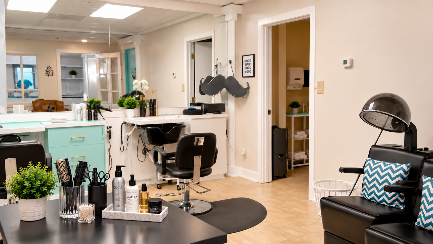 Interior of a hair salon with styling chairs, hair care products, and a reception area featuring black chairs with blue and white chevron pillows, a hair drying station, and wooden flooring.
