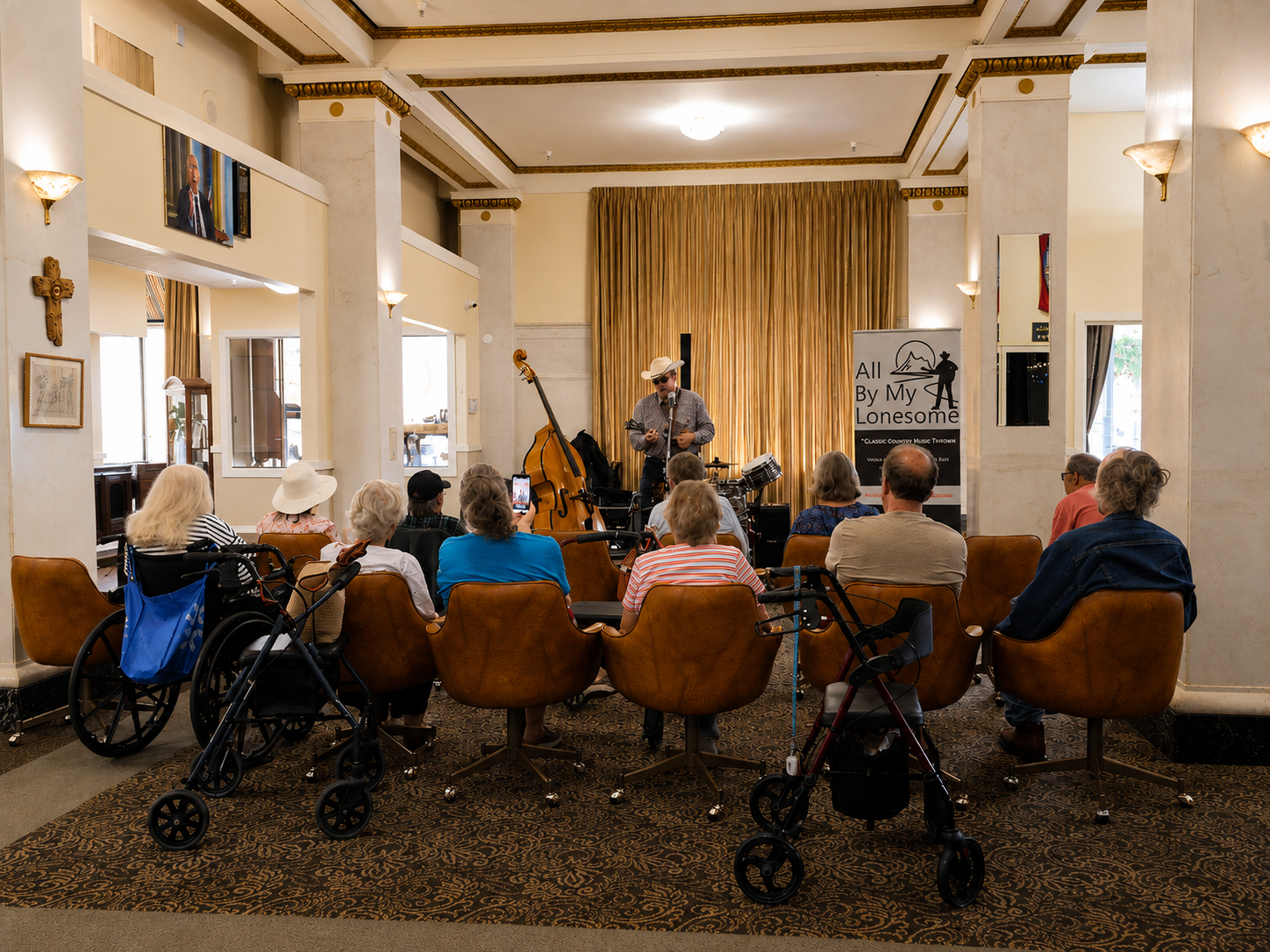 A small music performance with a singer and band on stage at an indoor venue, audience members seated in chairs, with some using walkers, and a banner that reads 'All By My Lonesome'.