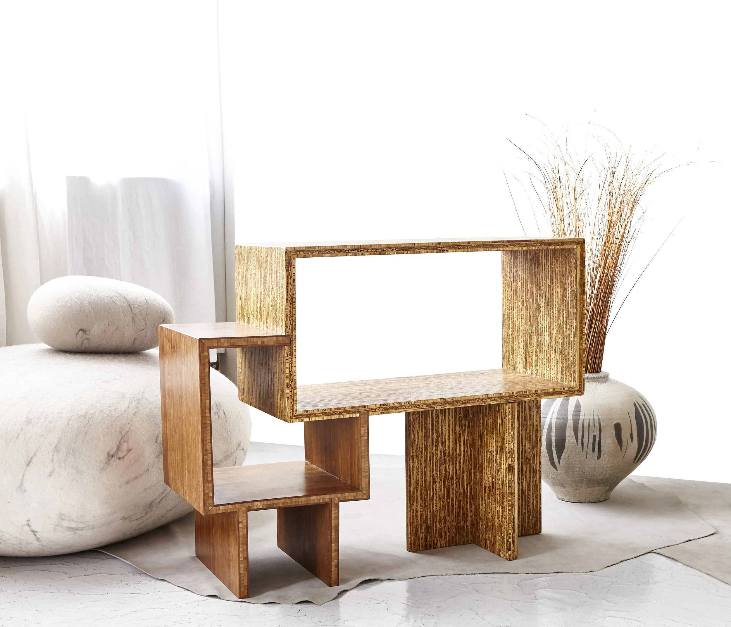 A wooden cube-shaped shelf with three open compartments in front of a white wall, accompanied by dried grass in a tall vase and a smaller rounded vase on the floor.