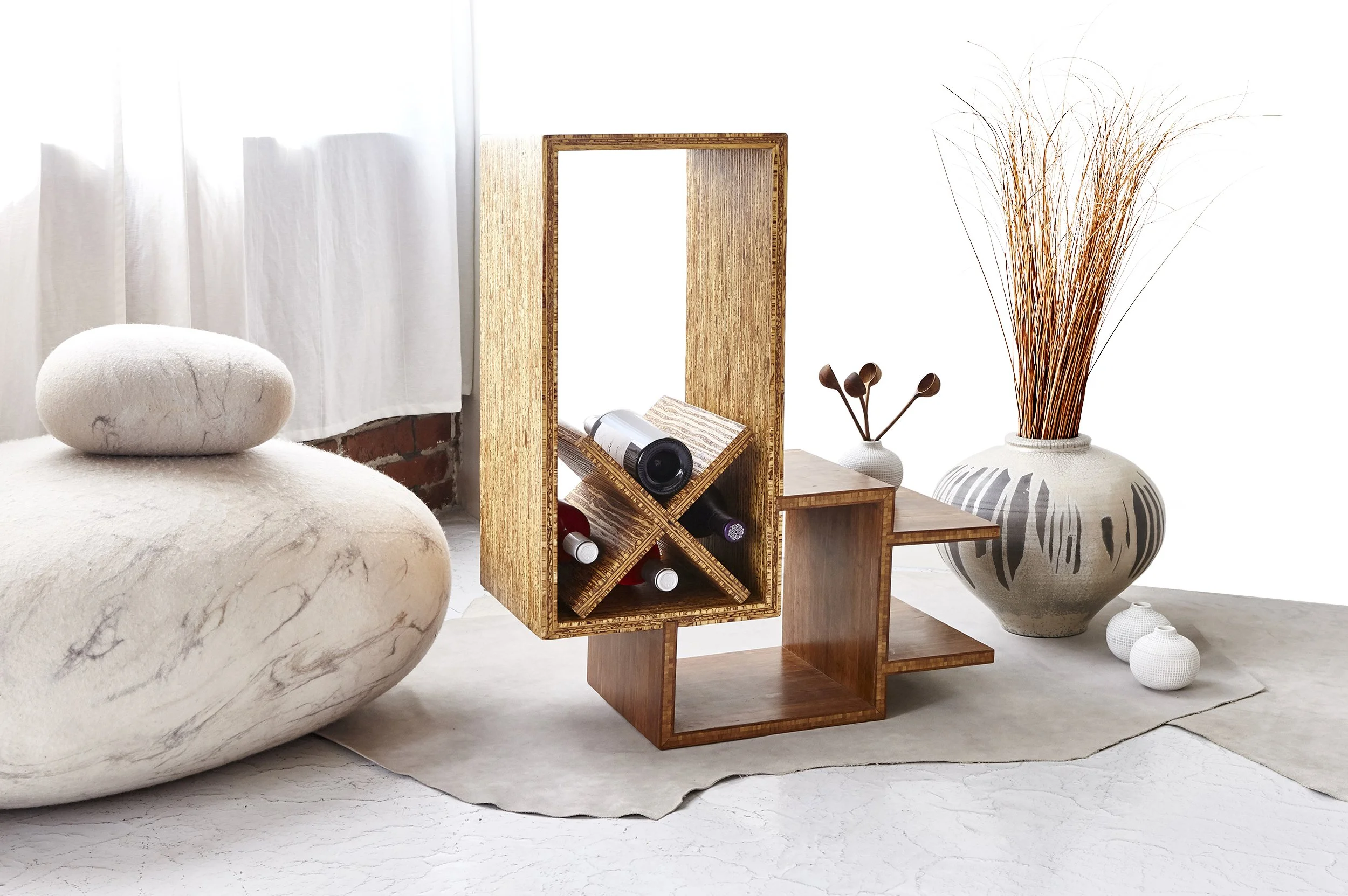 A modern living room corner with a round white marble ottoman, a tall wooden mirror, a small wooden side table with wine bottles, a large decorative vase with tall dried grass, and white curtains on a bright window.