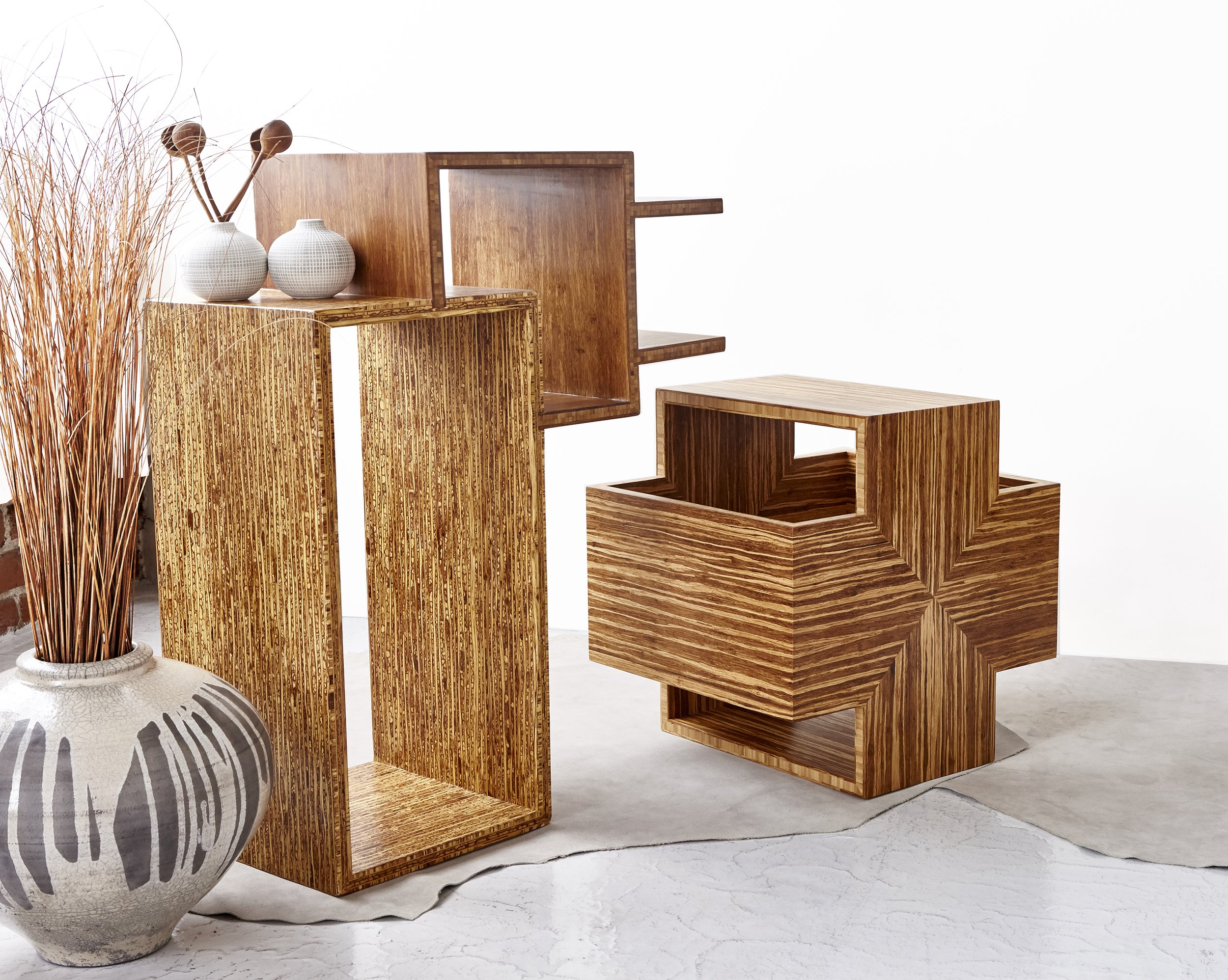 Group of wooden furniture pieces, including open shelves and tables, accompanied by a large white vase with dried reeds.
