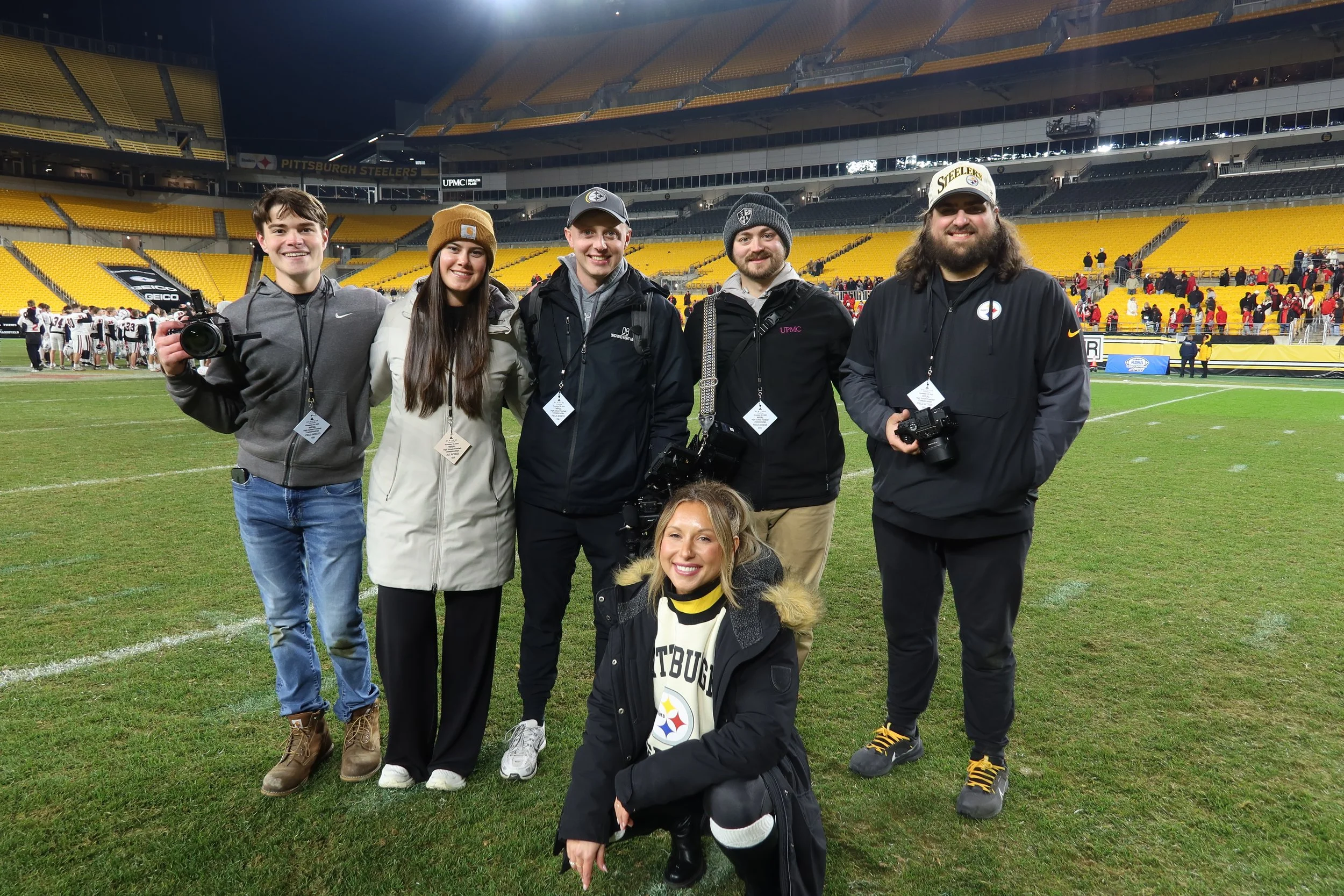Group of seven people, including five men and two women, standing on a football field at night with a stadium and football team in the background. The group is smiling and holding cameras, dressed warmly with some wearing Steelers apparel.