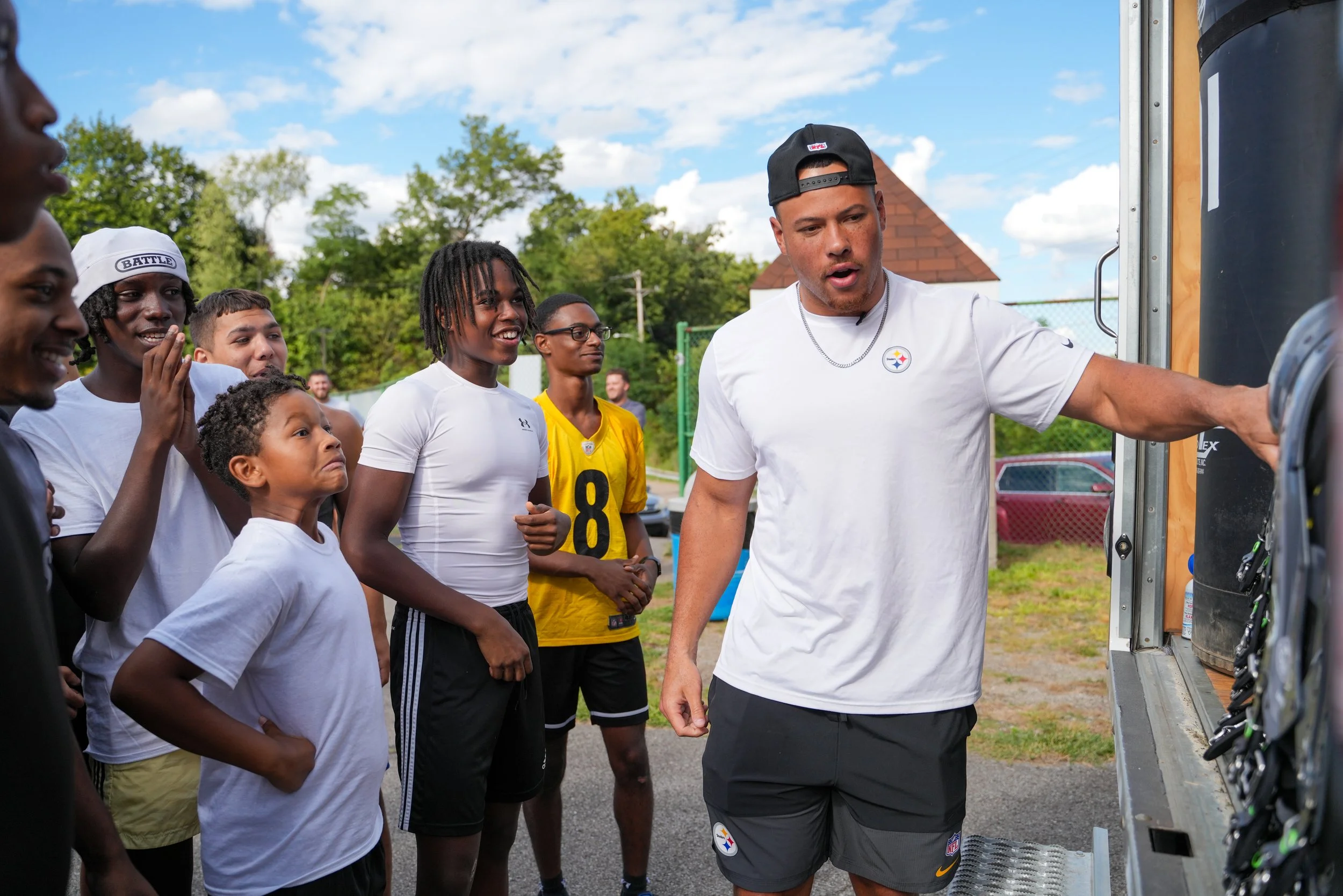 A group of young people, some wearing football jerseys, gathered around a man in athletic attire, watching him as he shows something on a vending machine or equipment.
