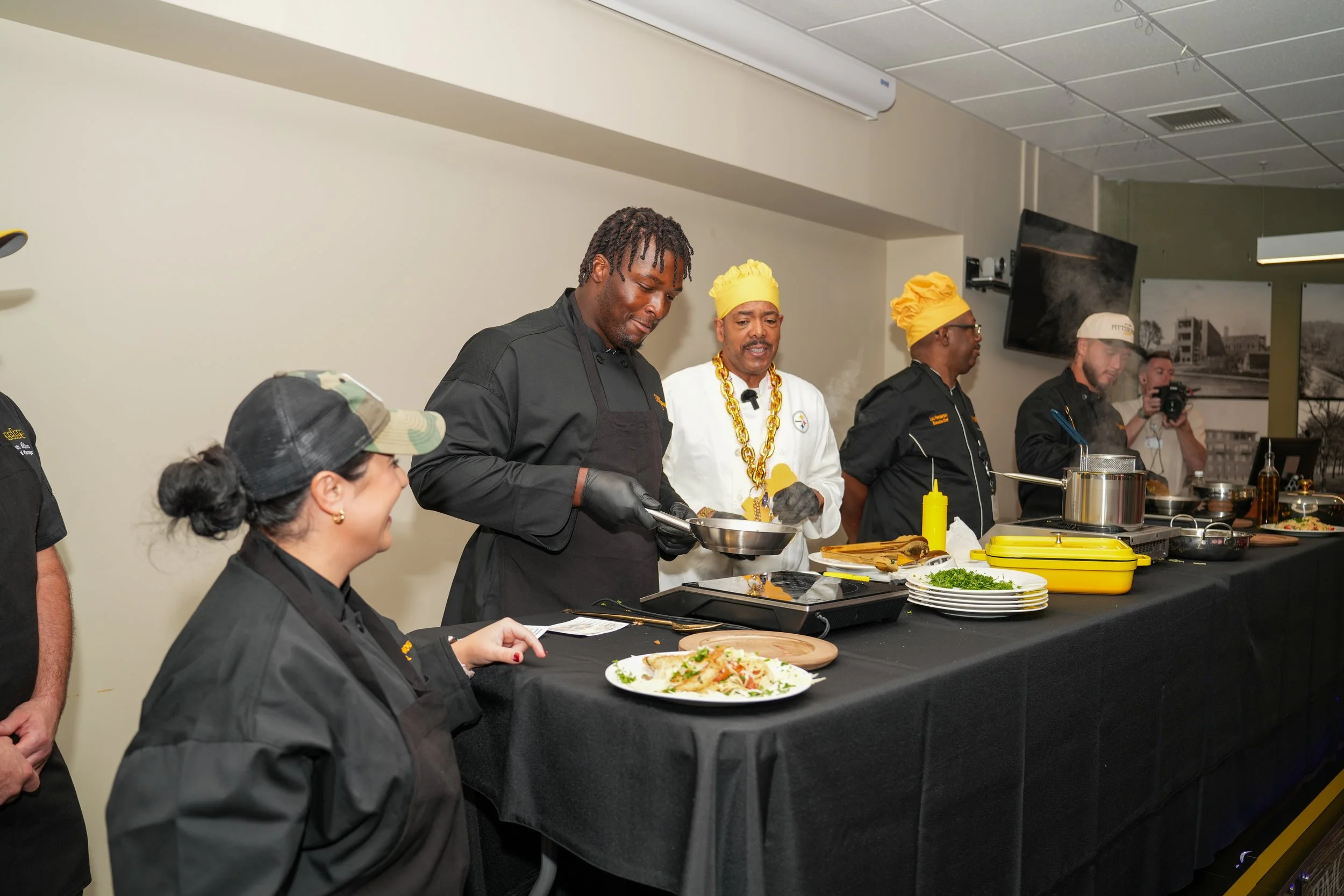 Group of chefs and a woman preparing food on a black table in a kitchen setting.