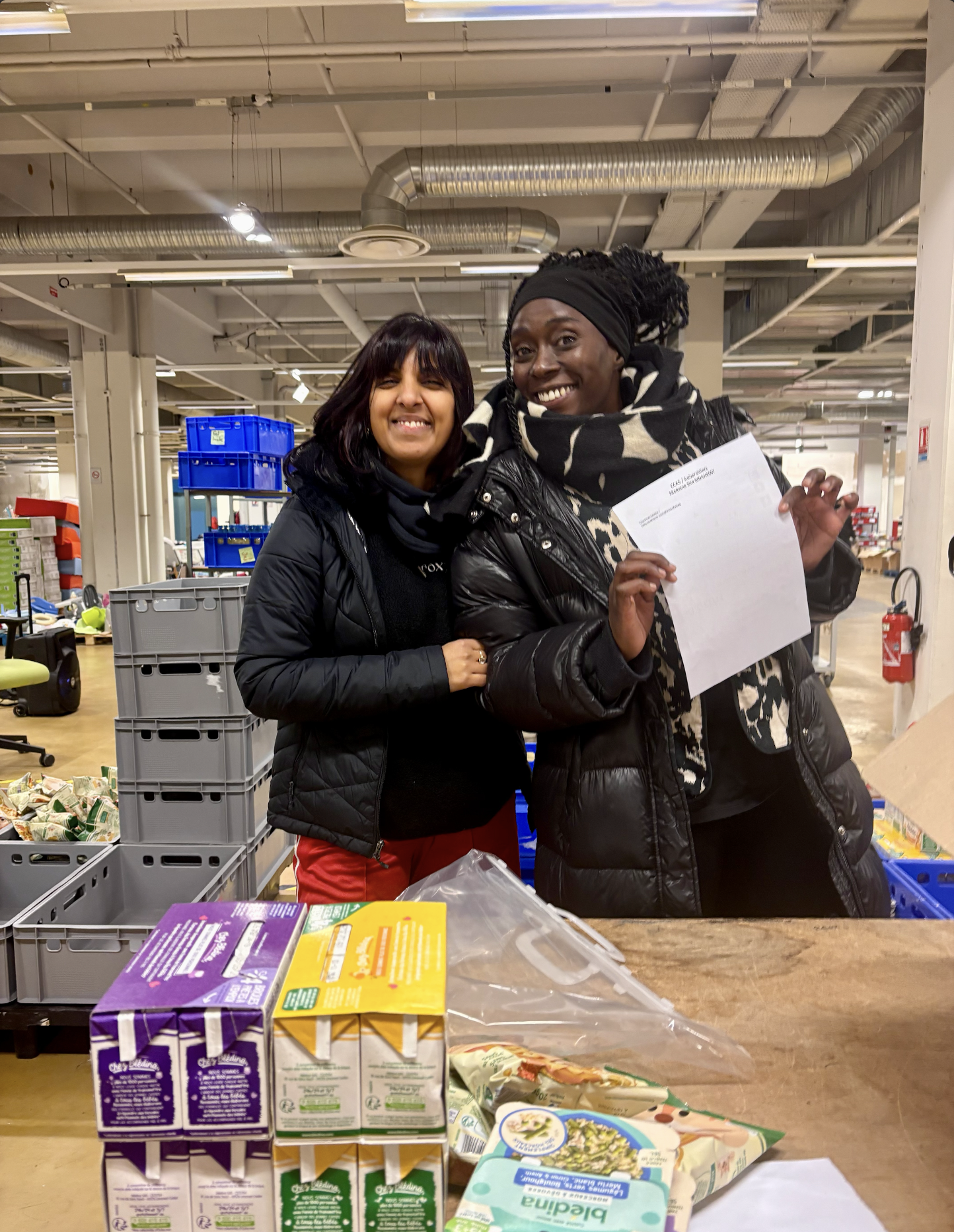 Deux femmes souriantes, portant des manteaux noirs, posent dans un entrepôt de supermarché, une d'elles tient un document.
