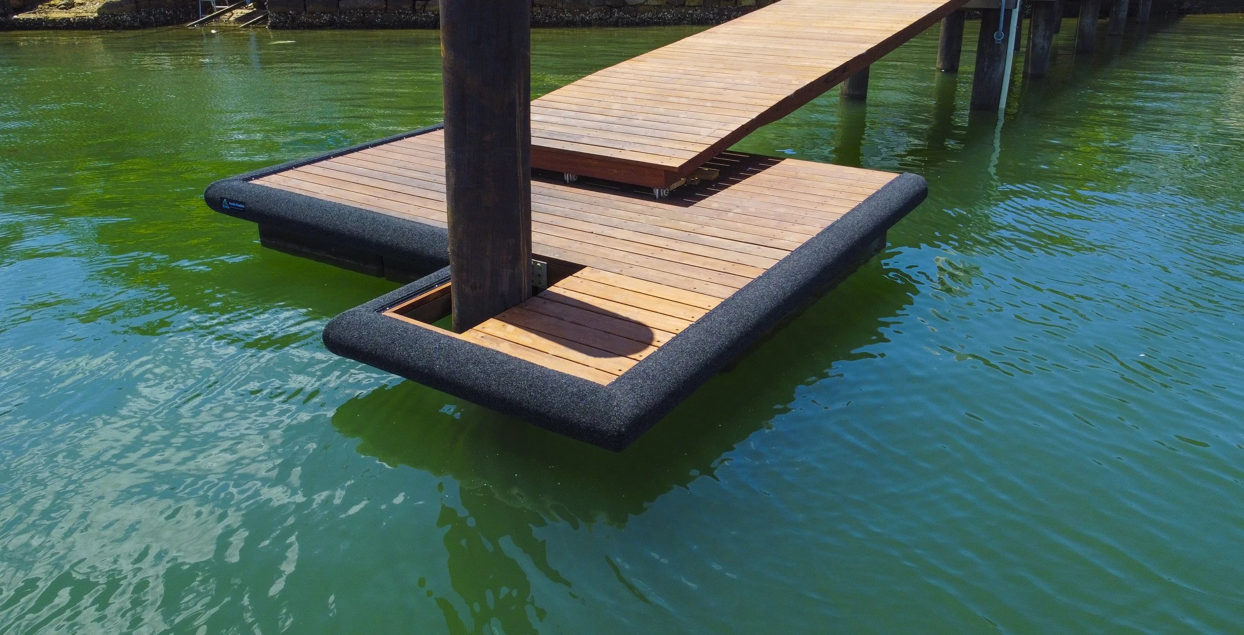 A wooden dock extending over green water with a partially submerged section that has black padding, supported by wooden pilings.