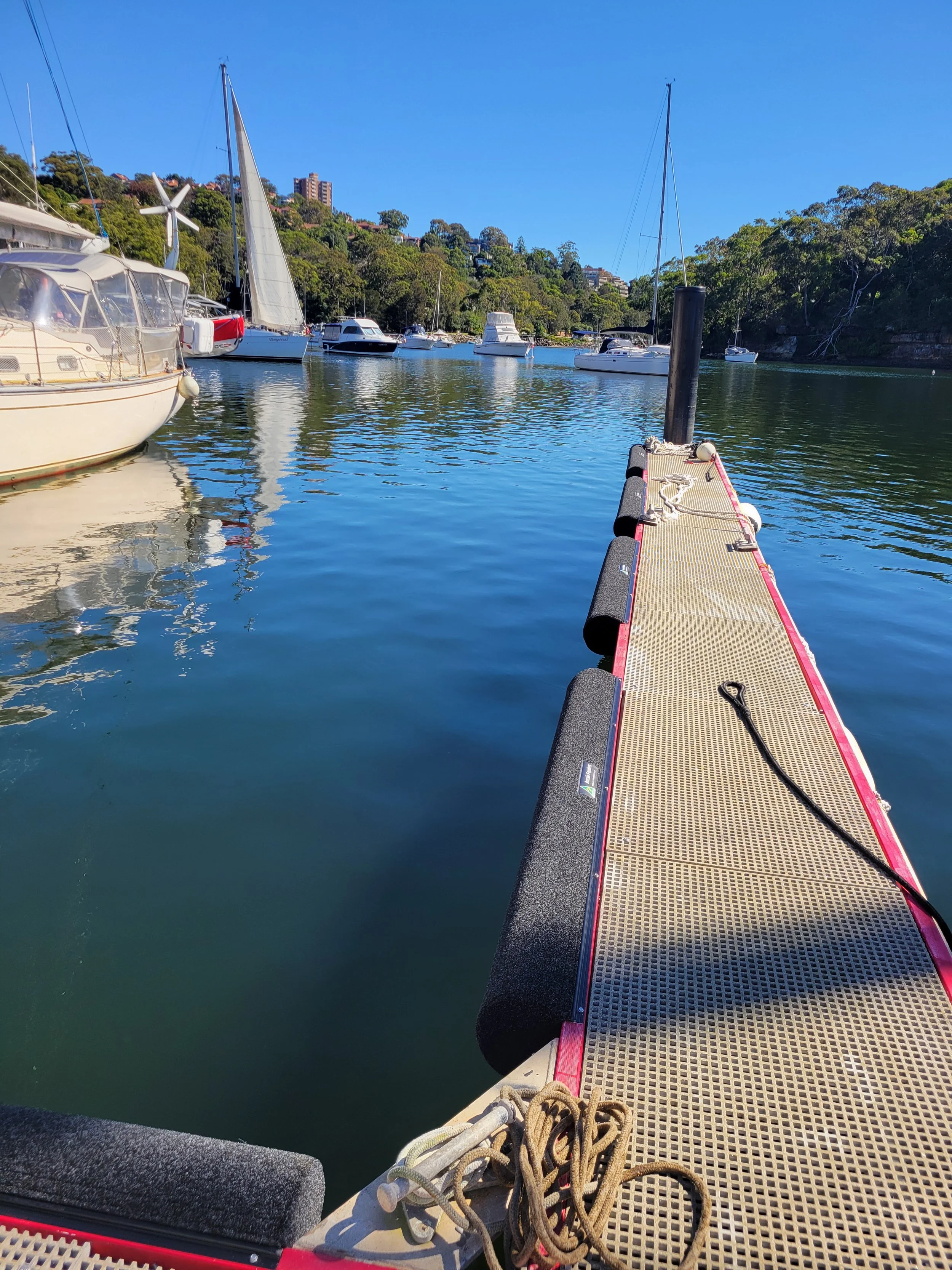 View from a boat dock showing a floating platform with ropes, nearby boats in the water, and a hillside with trees and buildings under a clear blue sky.
