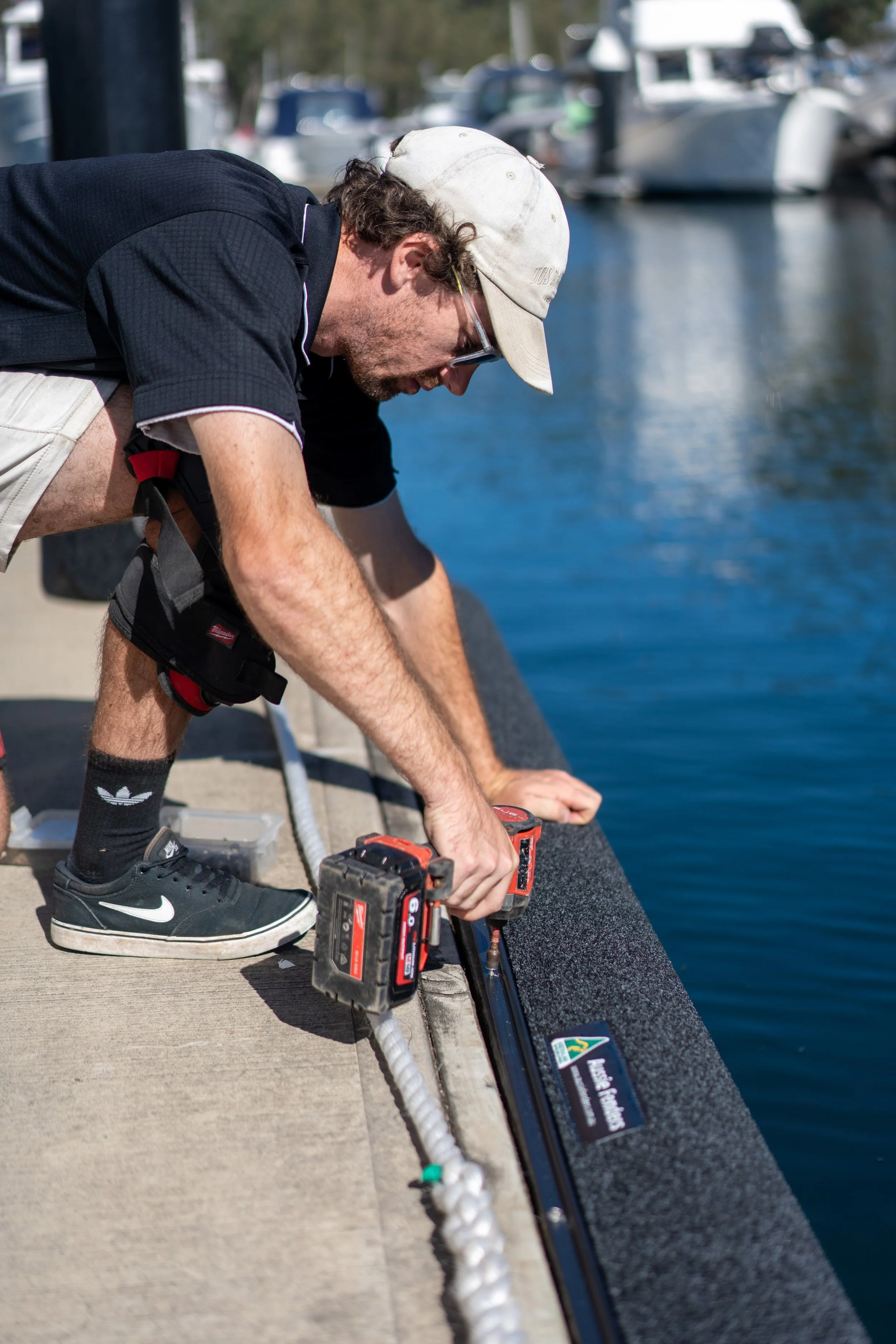 A tradesman using a power drill to install a black Aussie Fenders marine pontoon fender onto the edge of a concrete boat dock, with blue water and boats in the background.