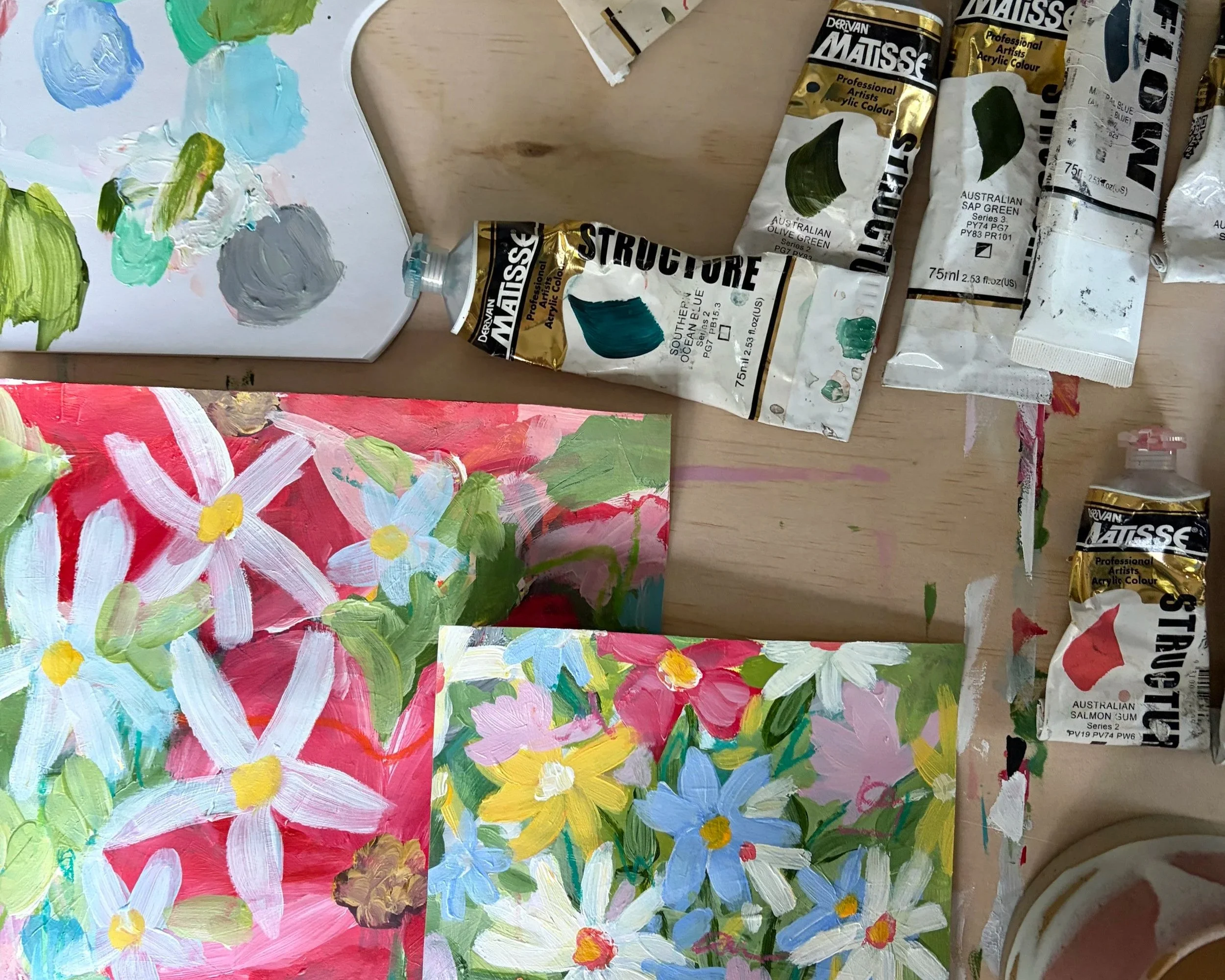 Colorful floral paintings on paper with visible paint tubes and brushes on a wooden table.