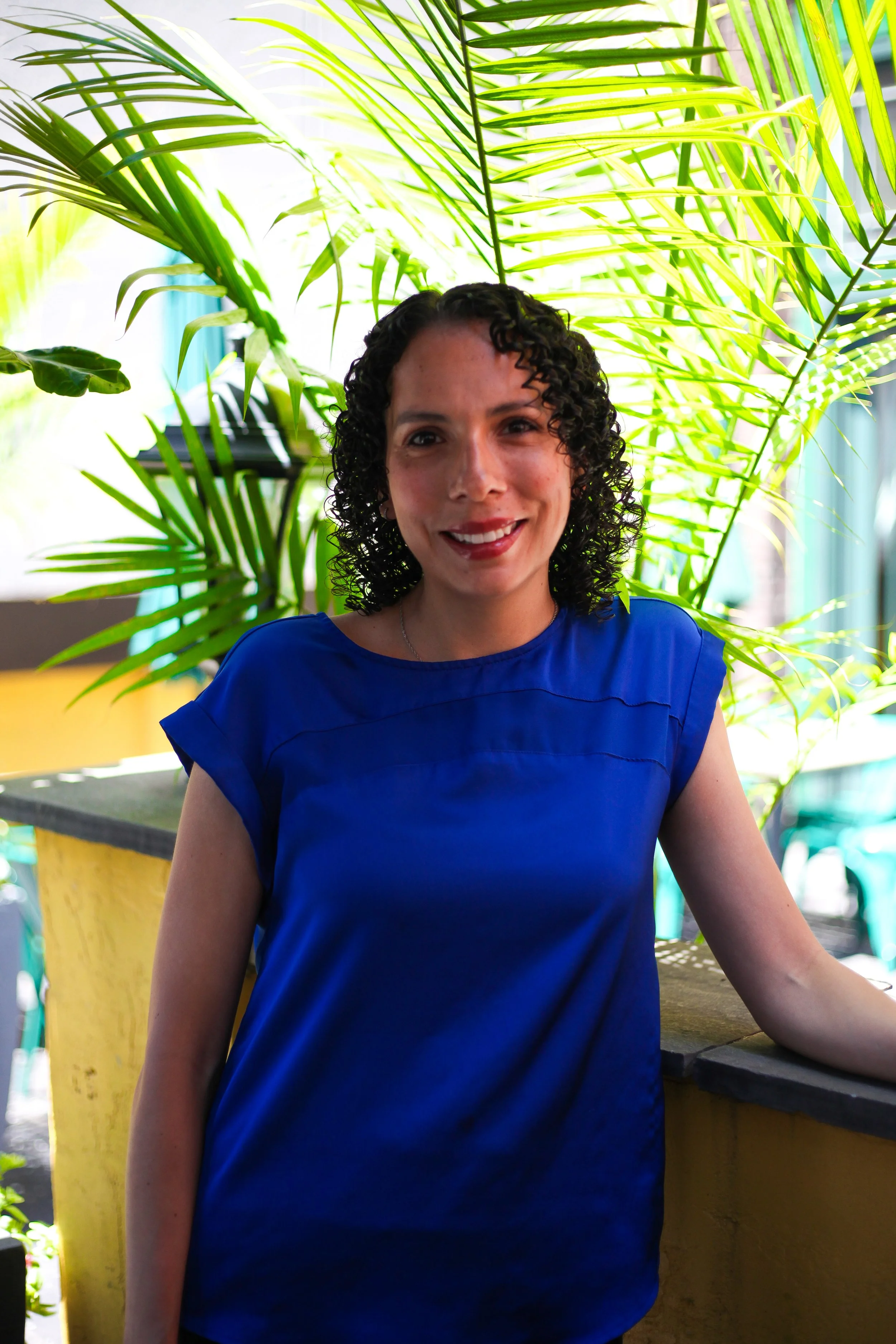A smiling woman in a royal blue shirt, standing outdoors with plants and sunshine in the background. 