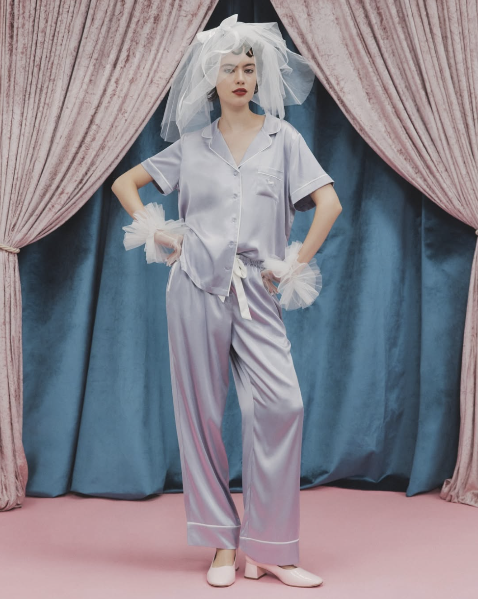 Woman in satin pajamas wearing a veil and tulle wrist cuffs, standing in front of pink and blue curtains.