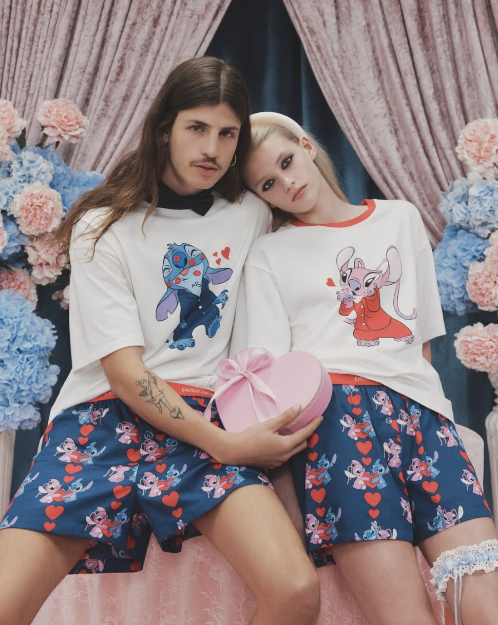A young man and woman sit close together, holding a pink heart-shaped box with a ribbon. They wear matching pajamas featuring characters from Disney's 'Lilo & Stitch,' with a backdrop of pink and blue flowers and pink curtains.