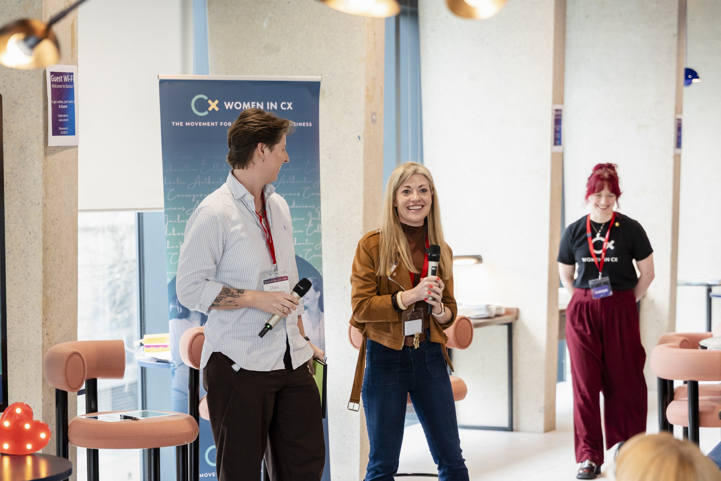 There is a lady in a brown jacket holding a microphone. Alongside her is a man in a blue shirt holding a microphone, with both appearing to be mid discussion. In the far right of the image, there is a lady walking towards the pair discussing.