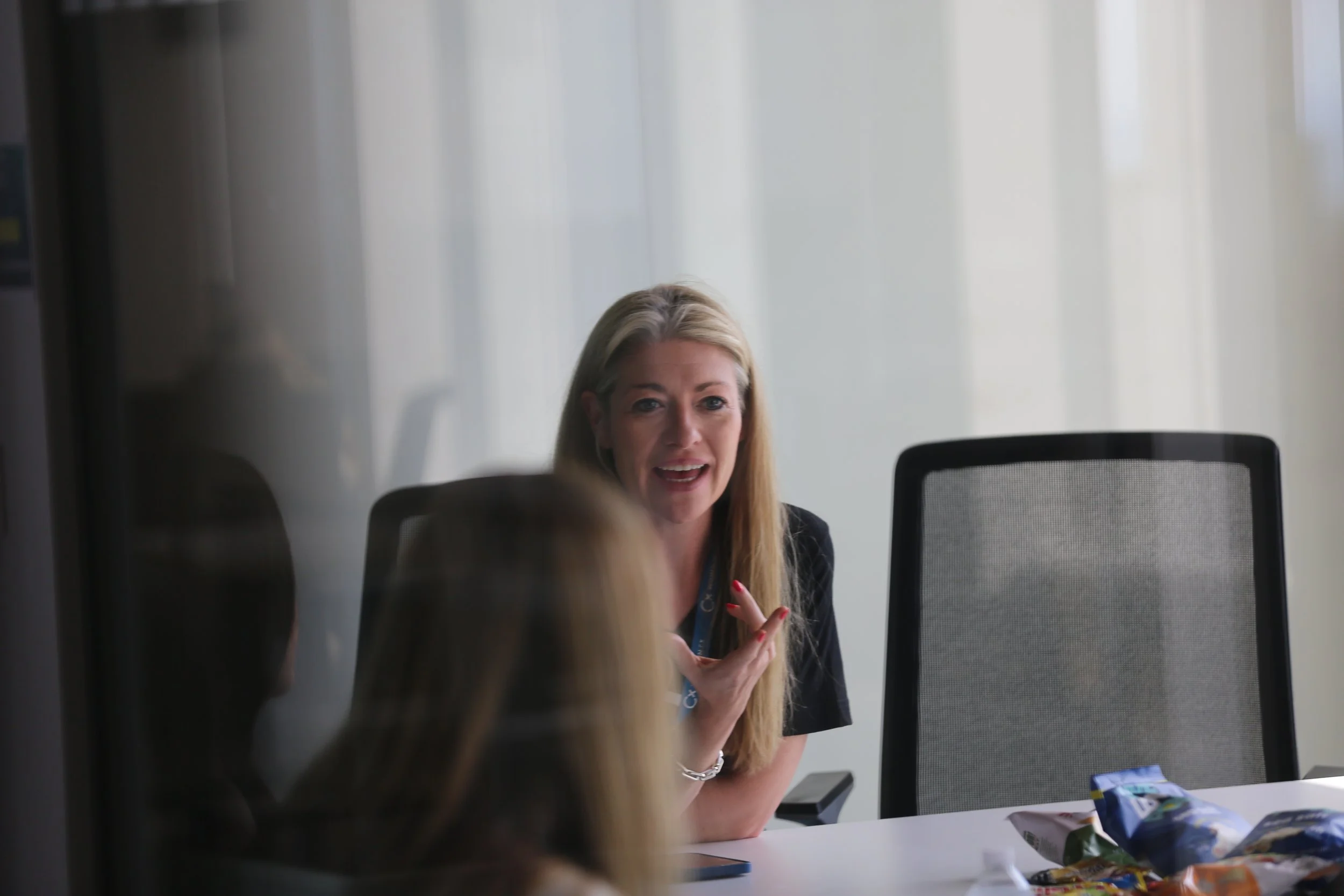 The image focuses on a woman who is mid conversation and talking with other individuals. there are black chairs they are sat on and the background is white. there is a blurred individuals head
