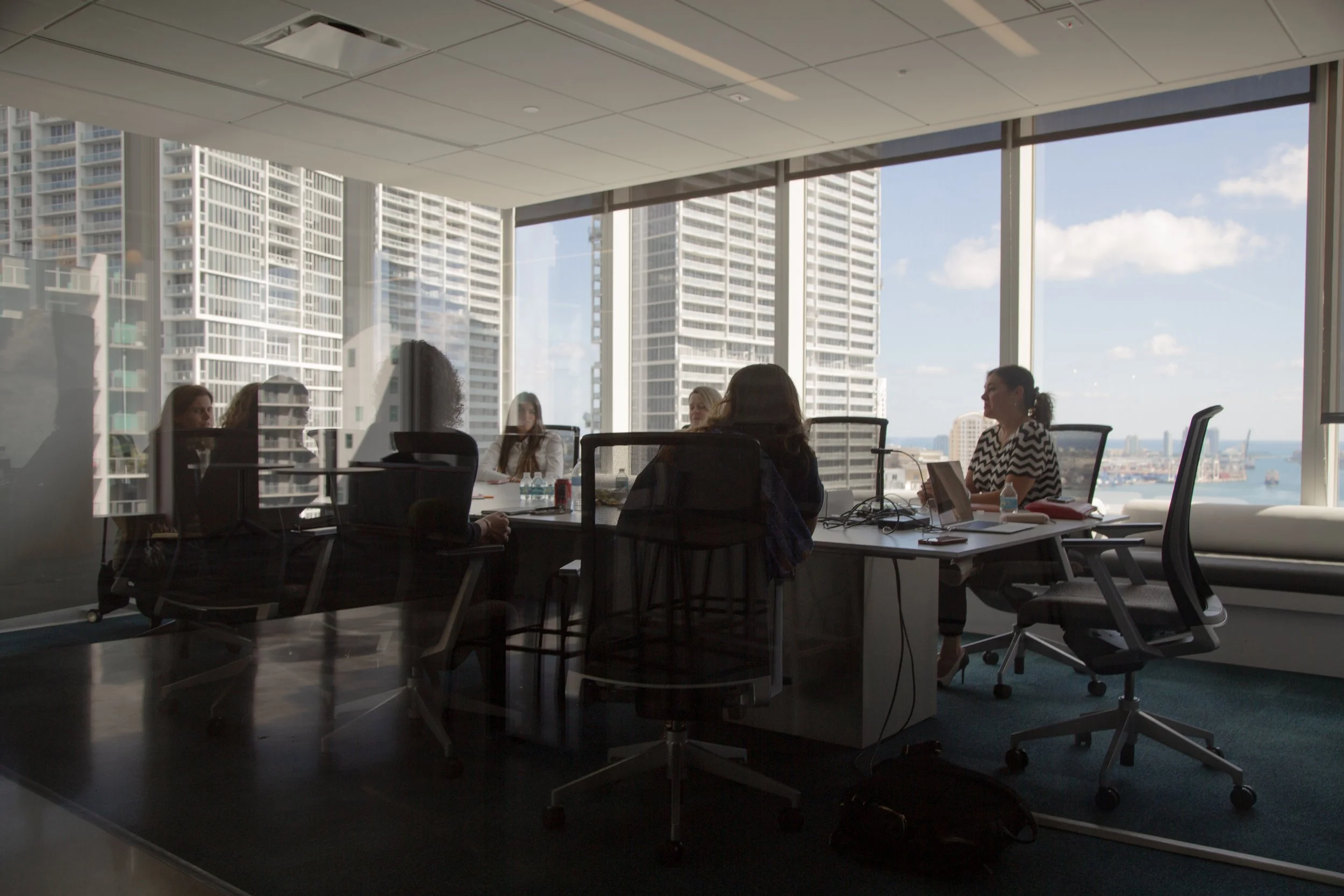Image of group of women sat around a large table in an office space with large glass windows, sky rise buildings visible in background.