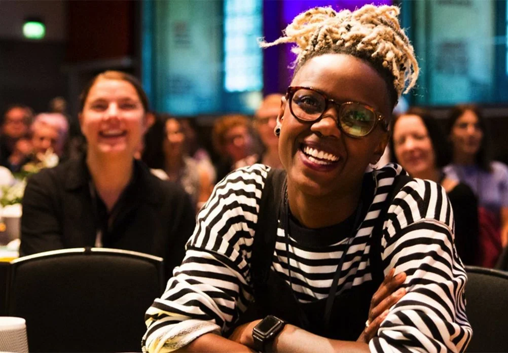 A young Black woman is smiling. She's wearing glasses, a black and white striped tee and black overalls. She's wearing a smart watch and glasses. Her hair is blonde, and in locs tied on top of her head. She looks happy.