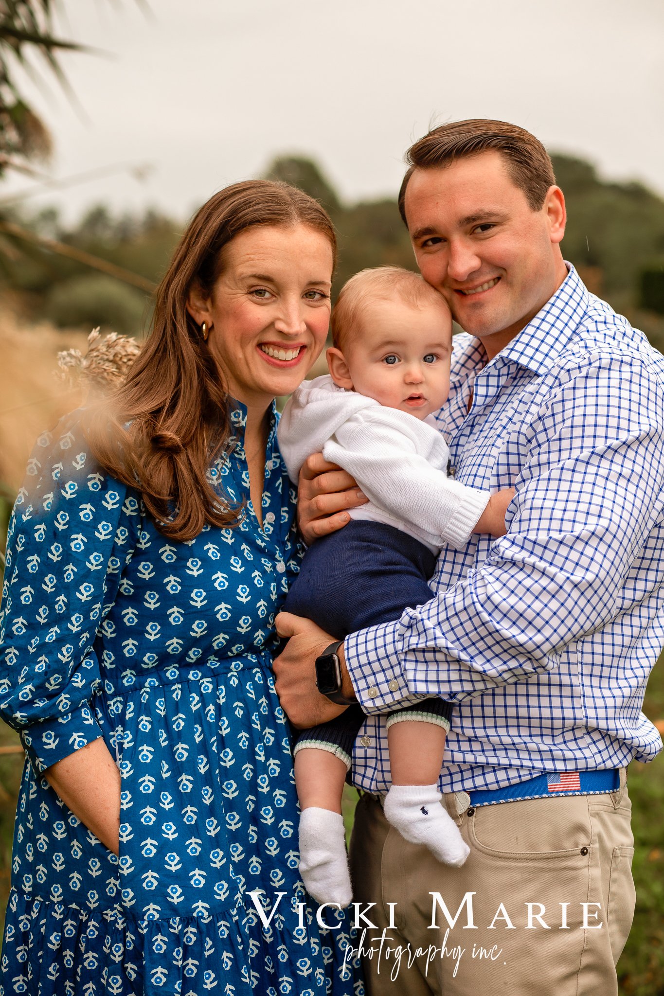 Family of three, mother, father, and baby, smiling outdoors on a cloudy day.