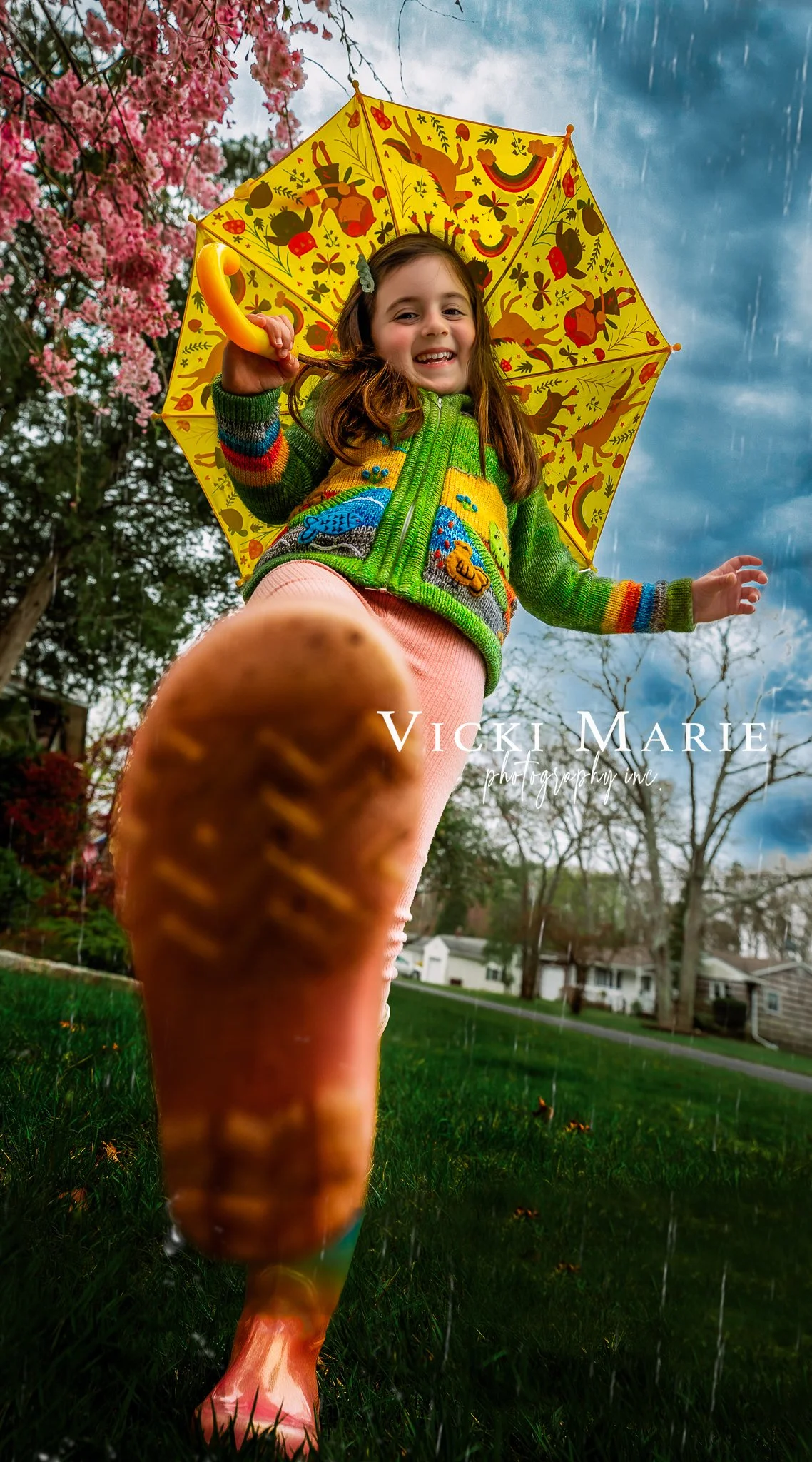 A young girl smiling, holding a yellow umbrella with colorful animal and nature patterns, dressed in a green sweater with animal designs and pink pants, standing outdoors on grass with trees and houses in the background.