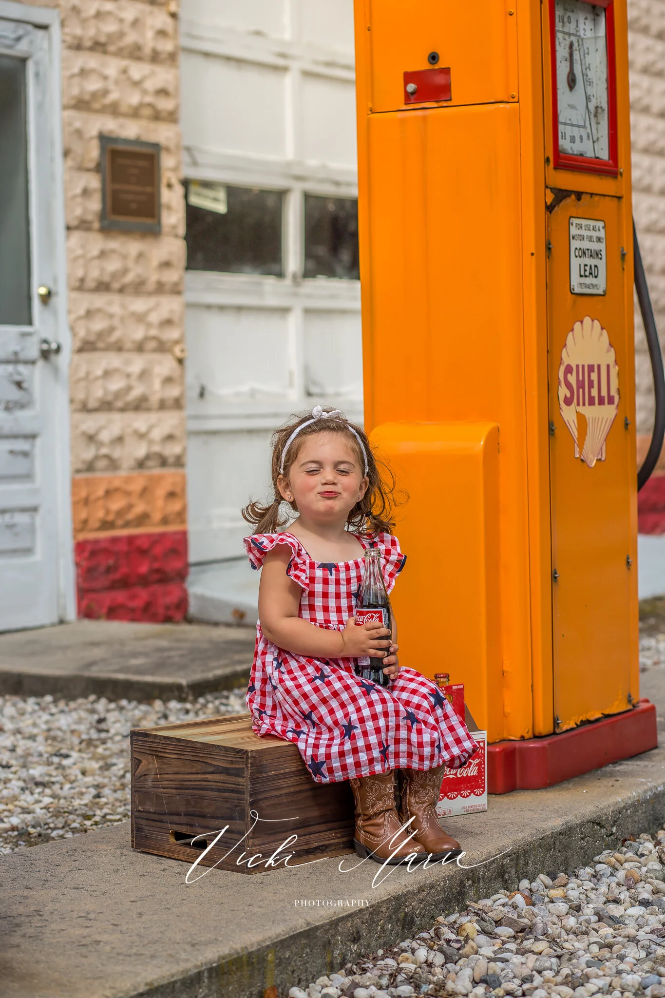 A young girl in a red and white checkered dress sitting on a wooden crate outside, holding a Coca-Cola bottle with her eyes closed and making a puckered face. An orange vintage gas pump with the Shell logo is next to her.