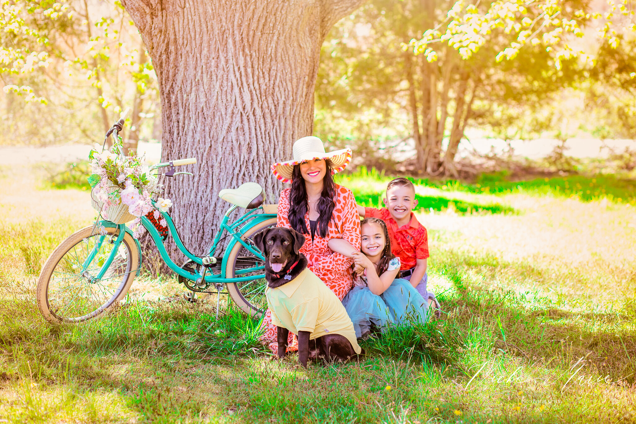 A woman with long black hair wearing a large sun hat and an orange dress, sitting on the grass with a black dog and two children, a girl and a boy, next to her. The woman and children are smiling. The girl has braided hair and is wearing a blue dress