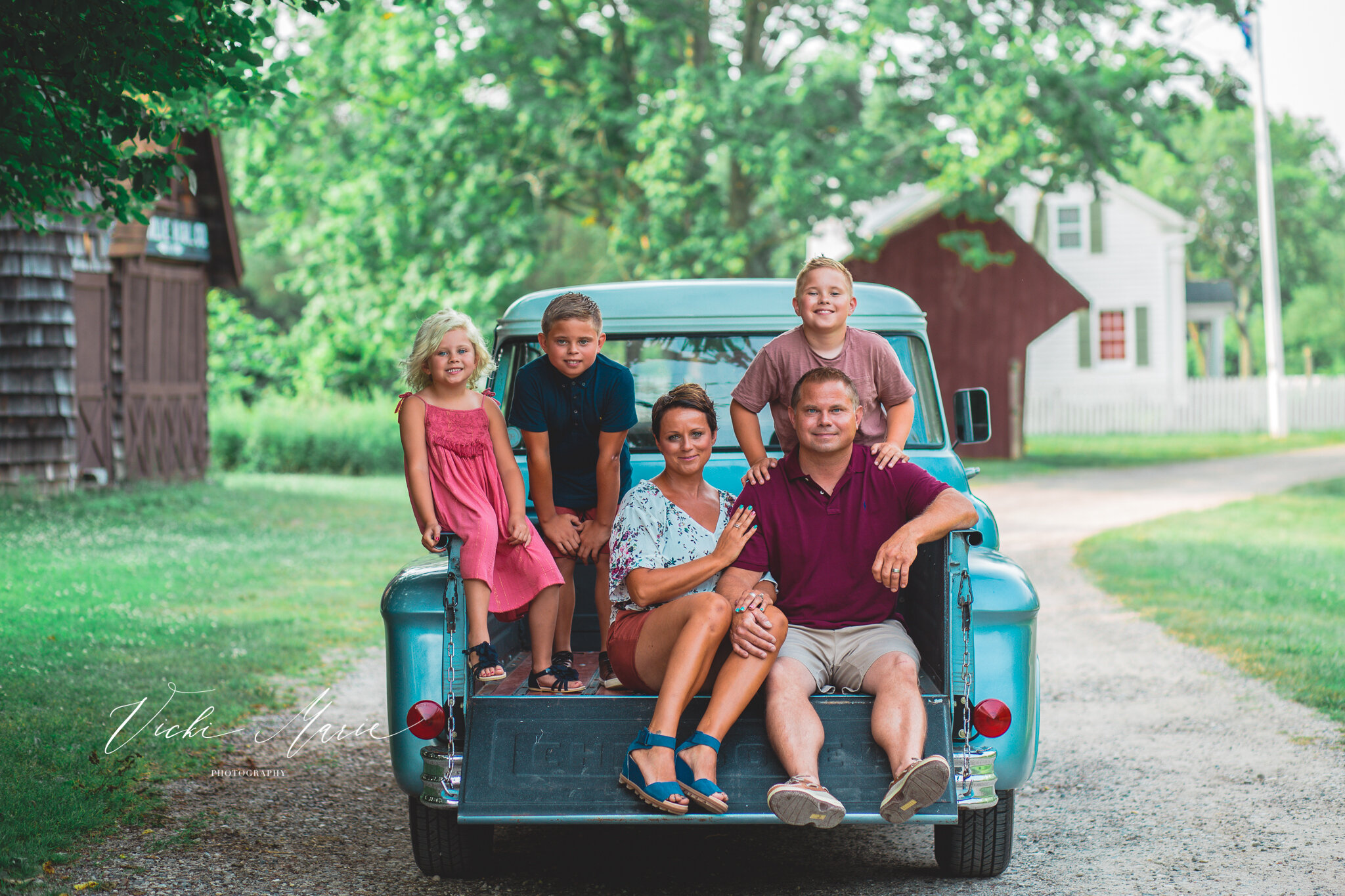 Family sitting on the back of a vintage blue truck outdoors, with green trees and a white house in the background.