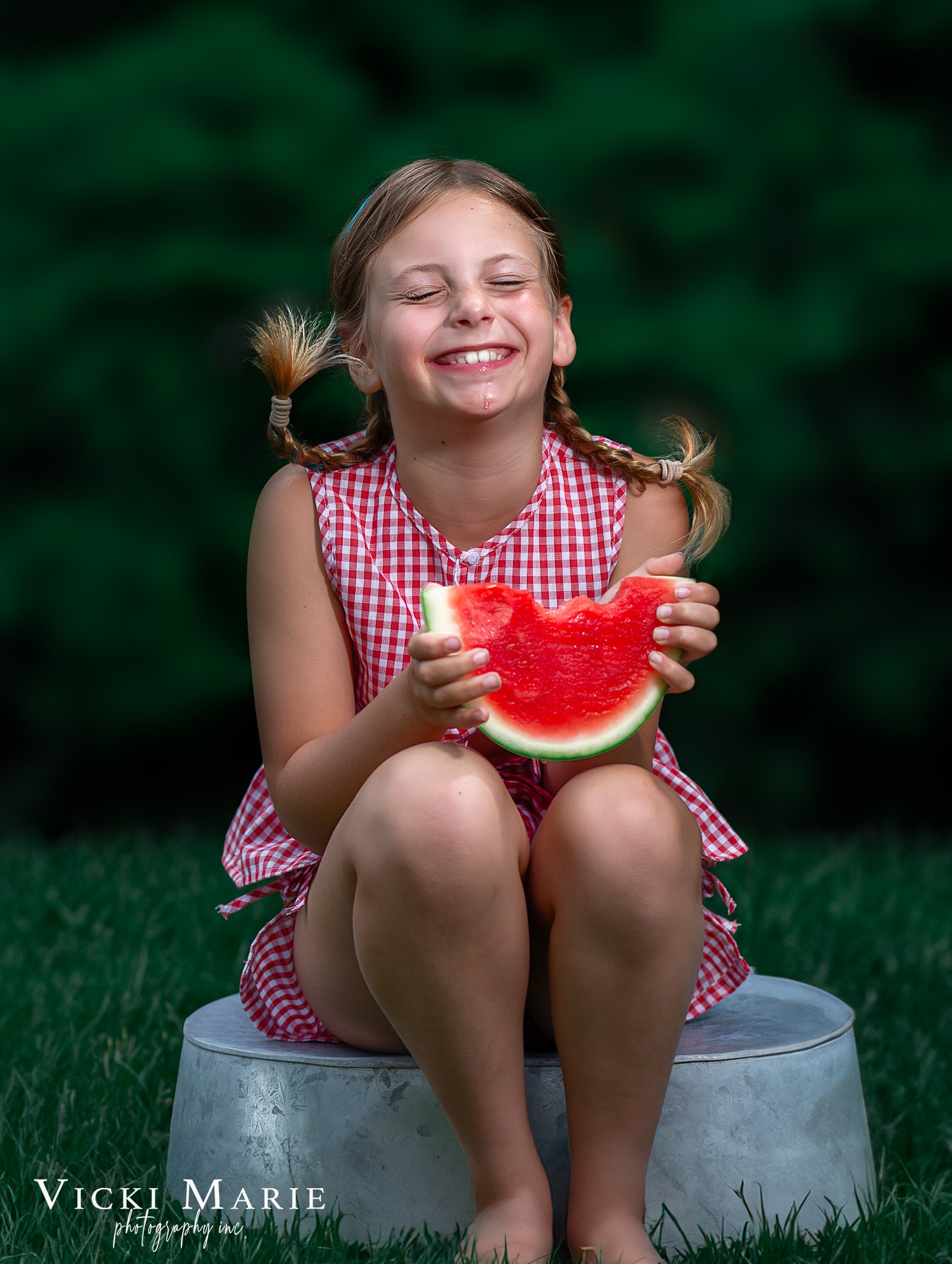 A young girl with braided hair and a red and white checkered dress sitting on a stool outdoors, smiling with her eyes closed, holding a large slice of watermelon.