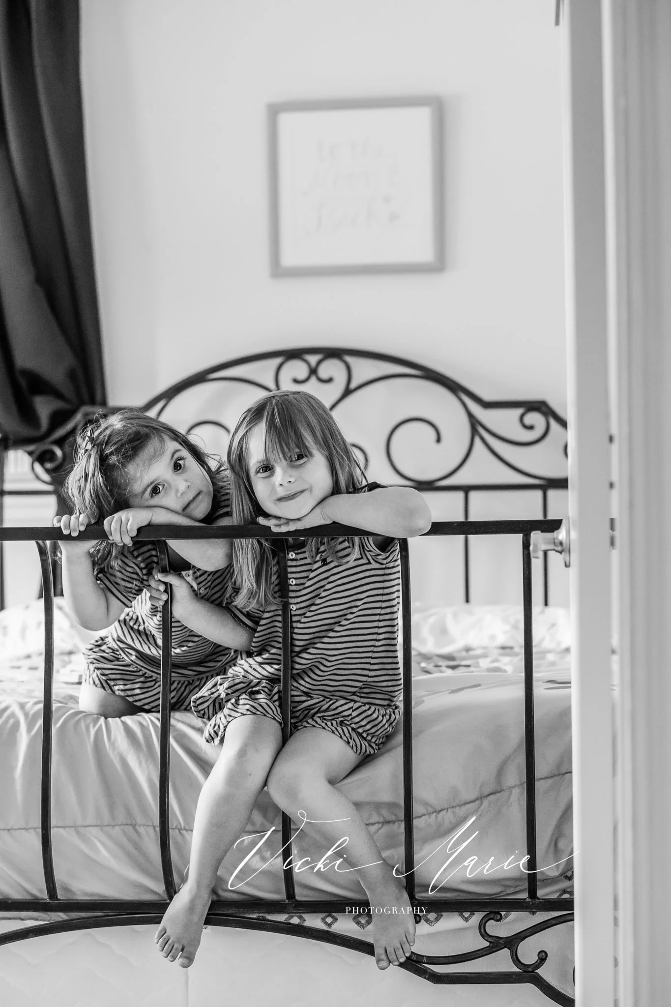 Two young girls in pajamas sitting on a bed, leaning over the iron headboard, smiling at the camera.