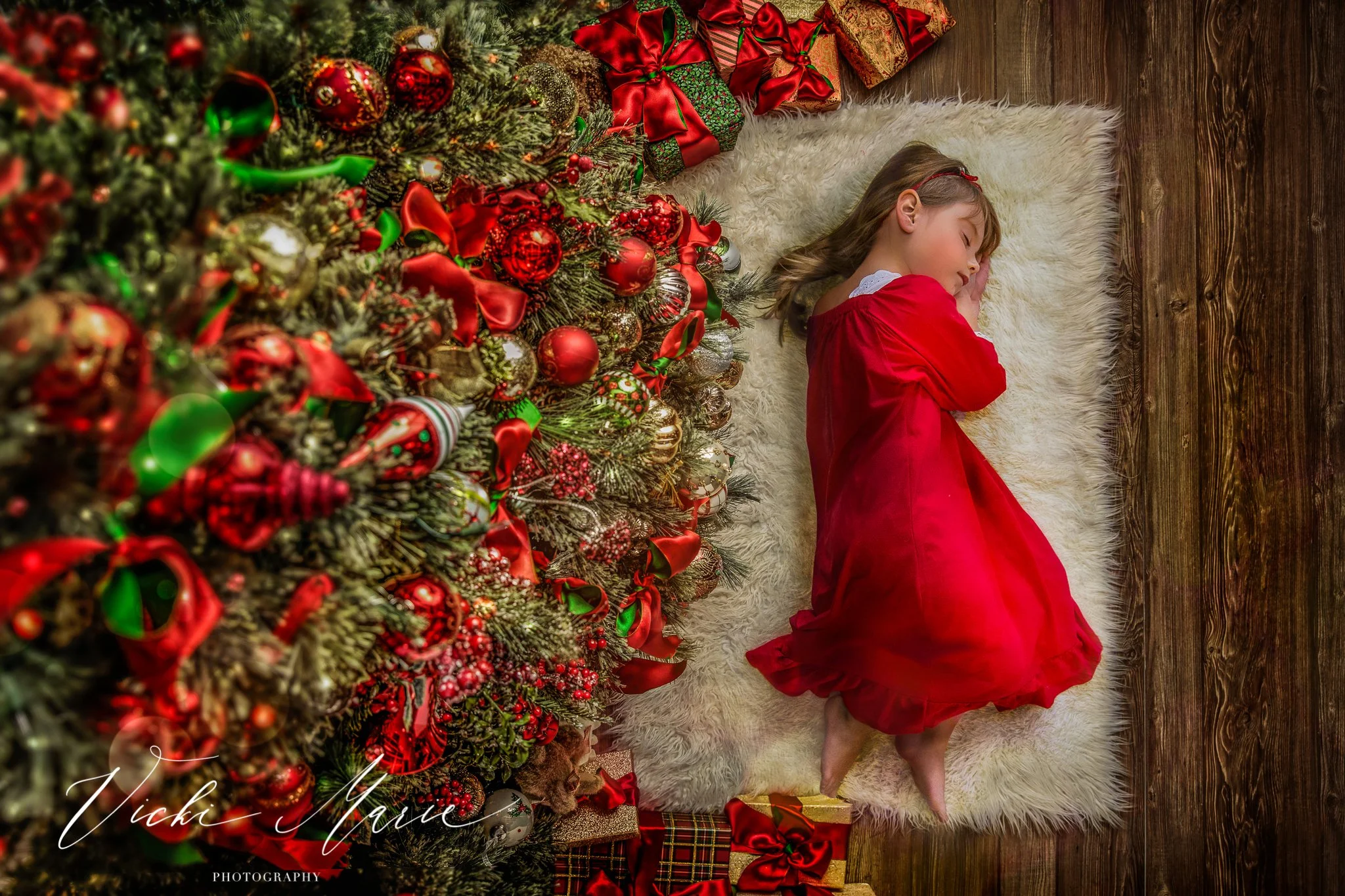 A young girl in a red dress is sleeping on a fluffy white rug next to a decorated Christmas tree with red, green, and gold ornaments. There are Christmas presents wrapped in colorful paper with ribbons underneath the tree.
