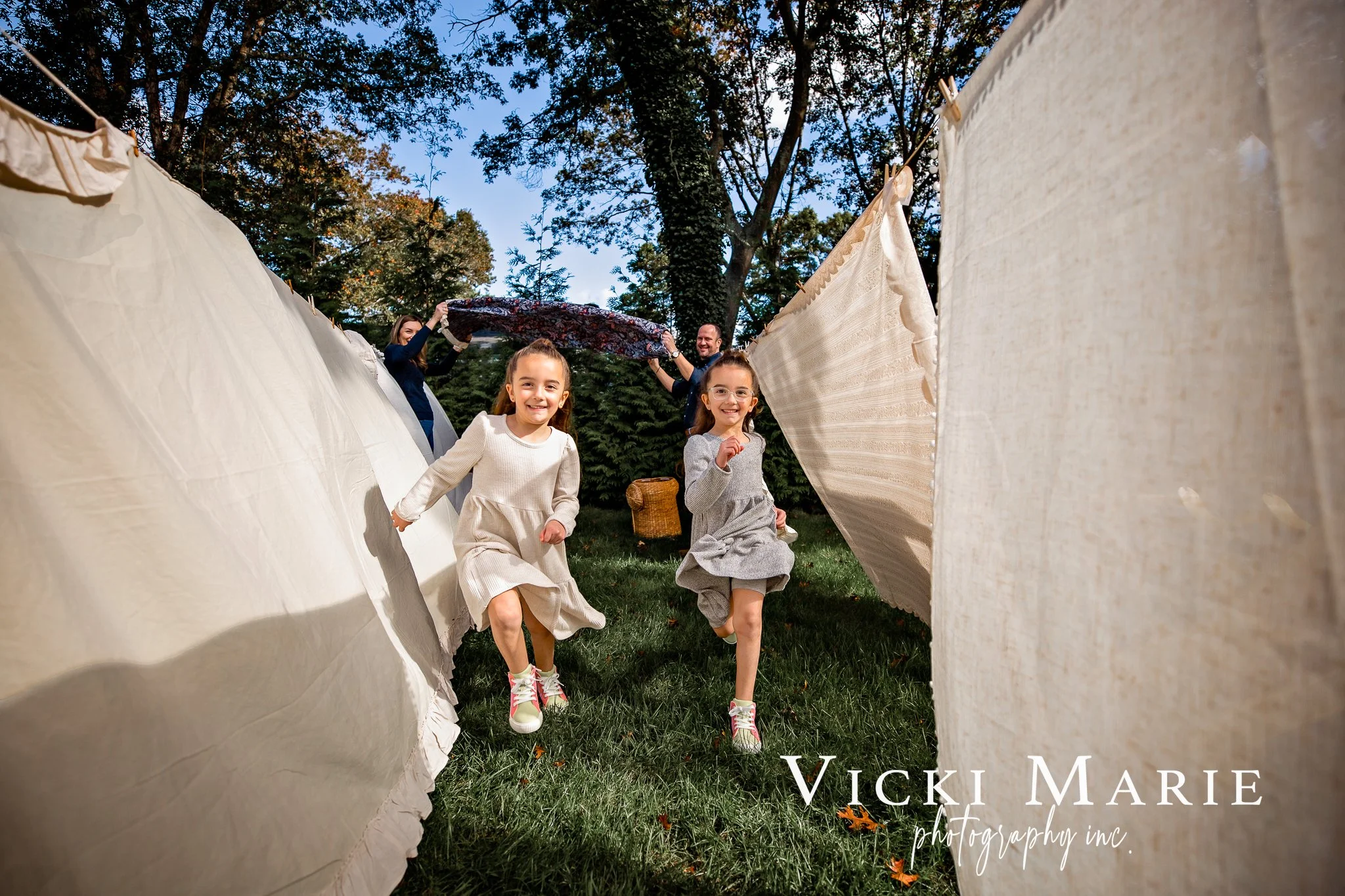 Two young girls in dresses running through a clothesline with hanging linens, with two adults hanging a blanket in the background in a park surrounded by trees.