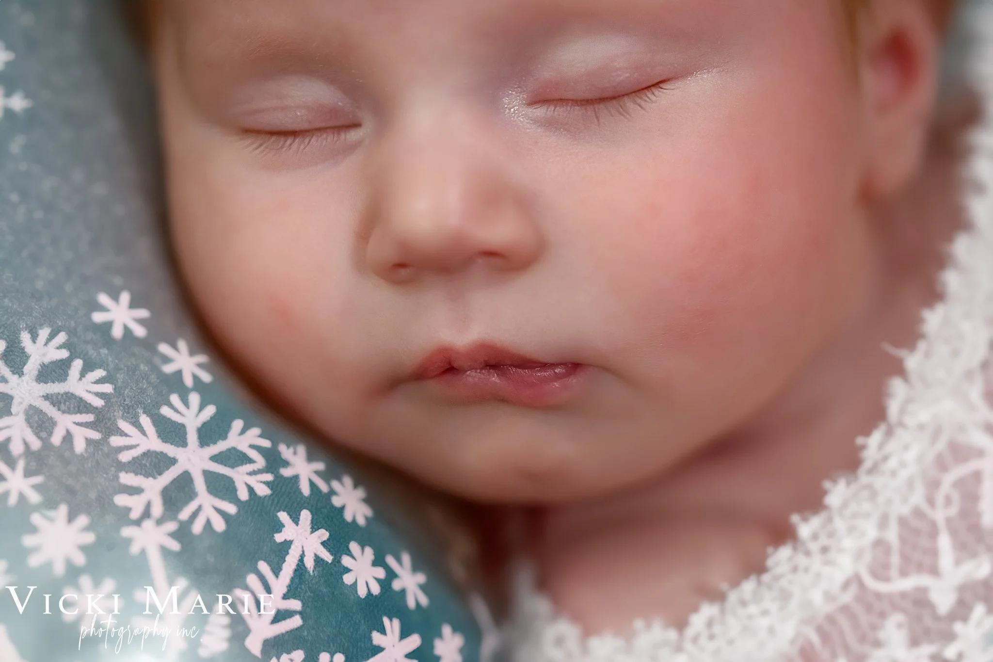 Close-up of a sleeping baby with closed eyes, resting on a pillow with snowflake pattern, wearing a lacy outfit.