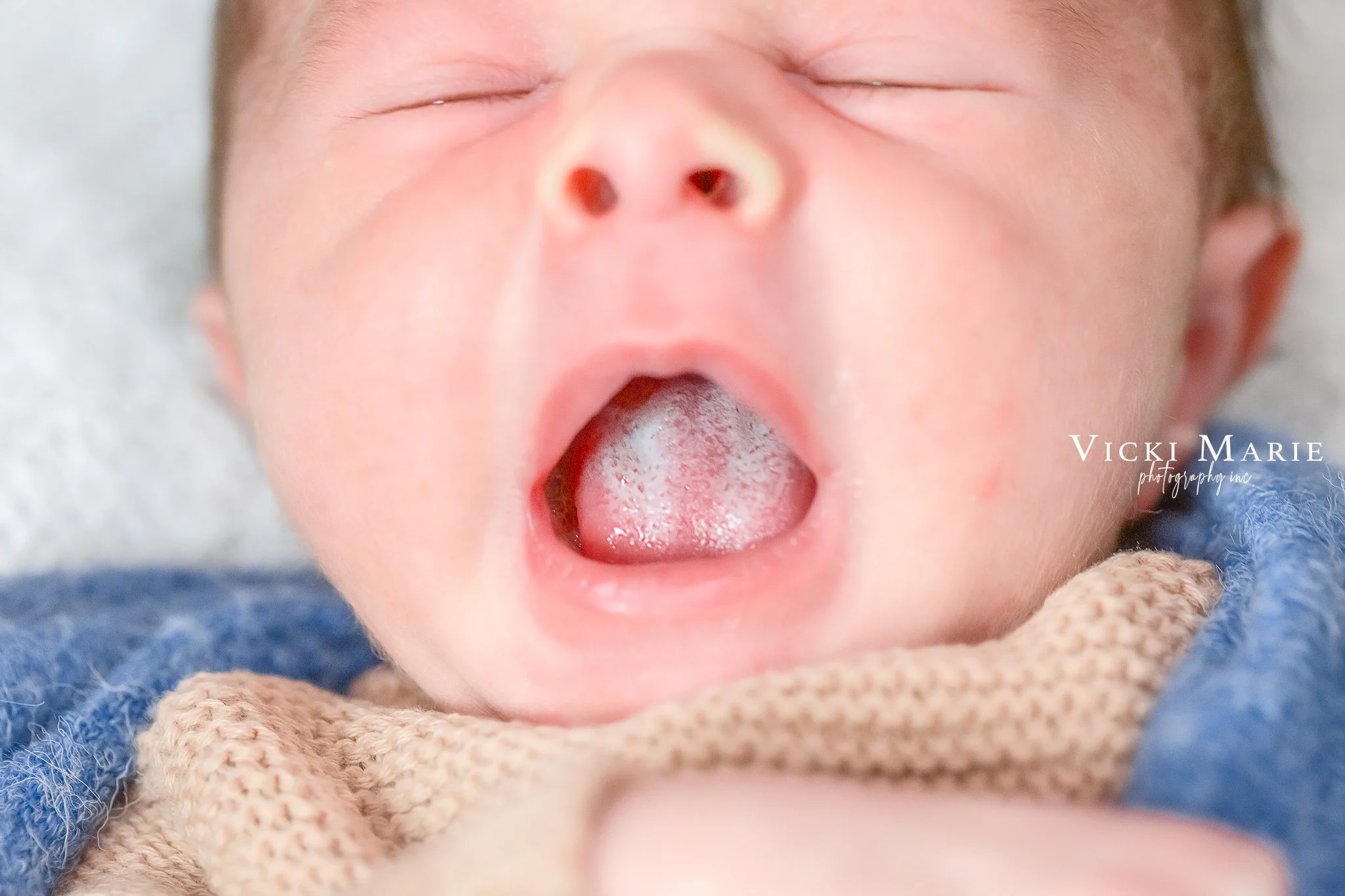 Close-up of a baby's face with mouth wide open, tongue visible, and eyes closed, lying on a beige and blue blanket.