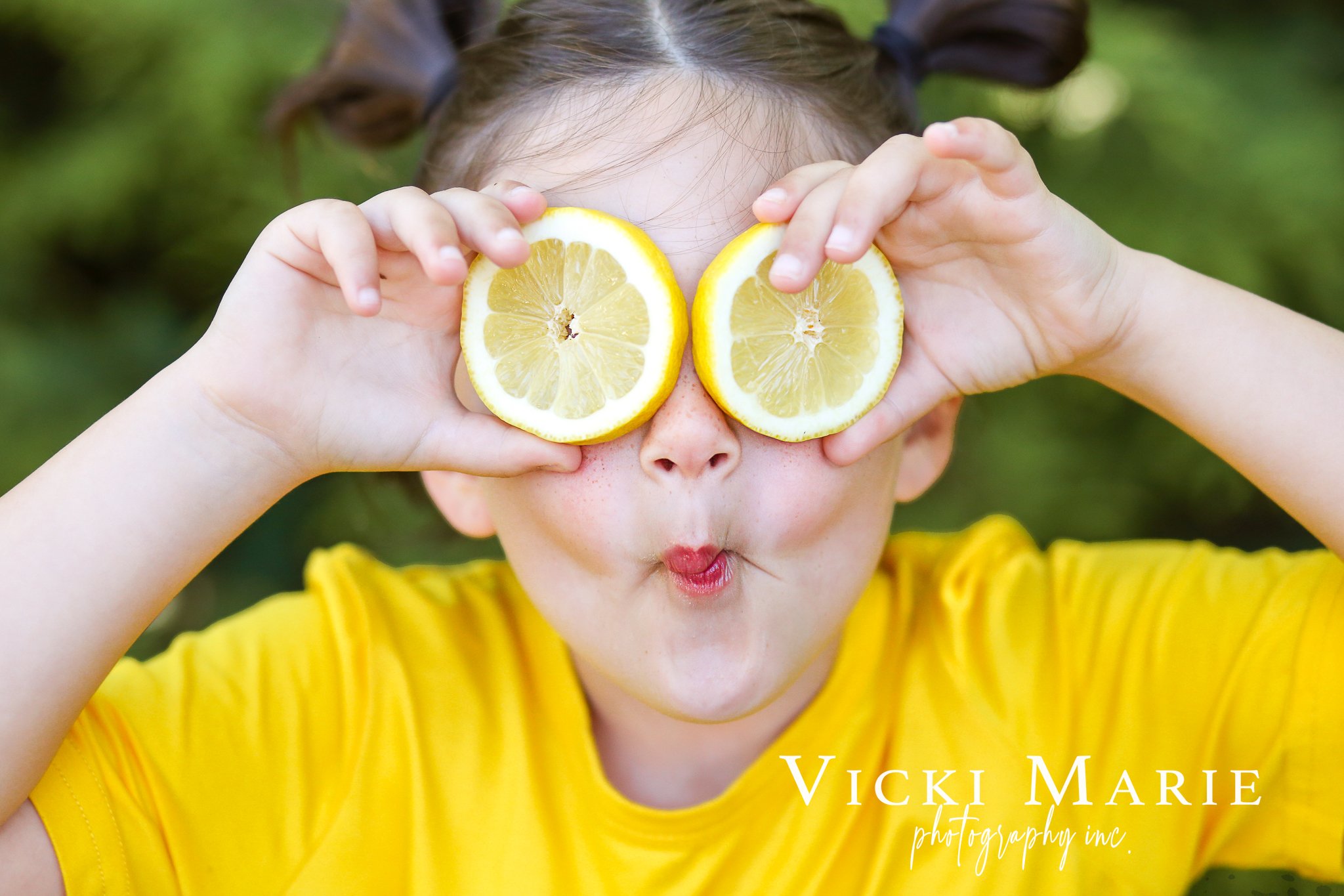 A young girl with pigtails holds two lemon halves over her eyes, making a playful face with puckered lips, wearing a yellow shirt, with a blurred green background.