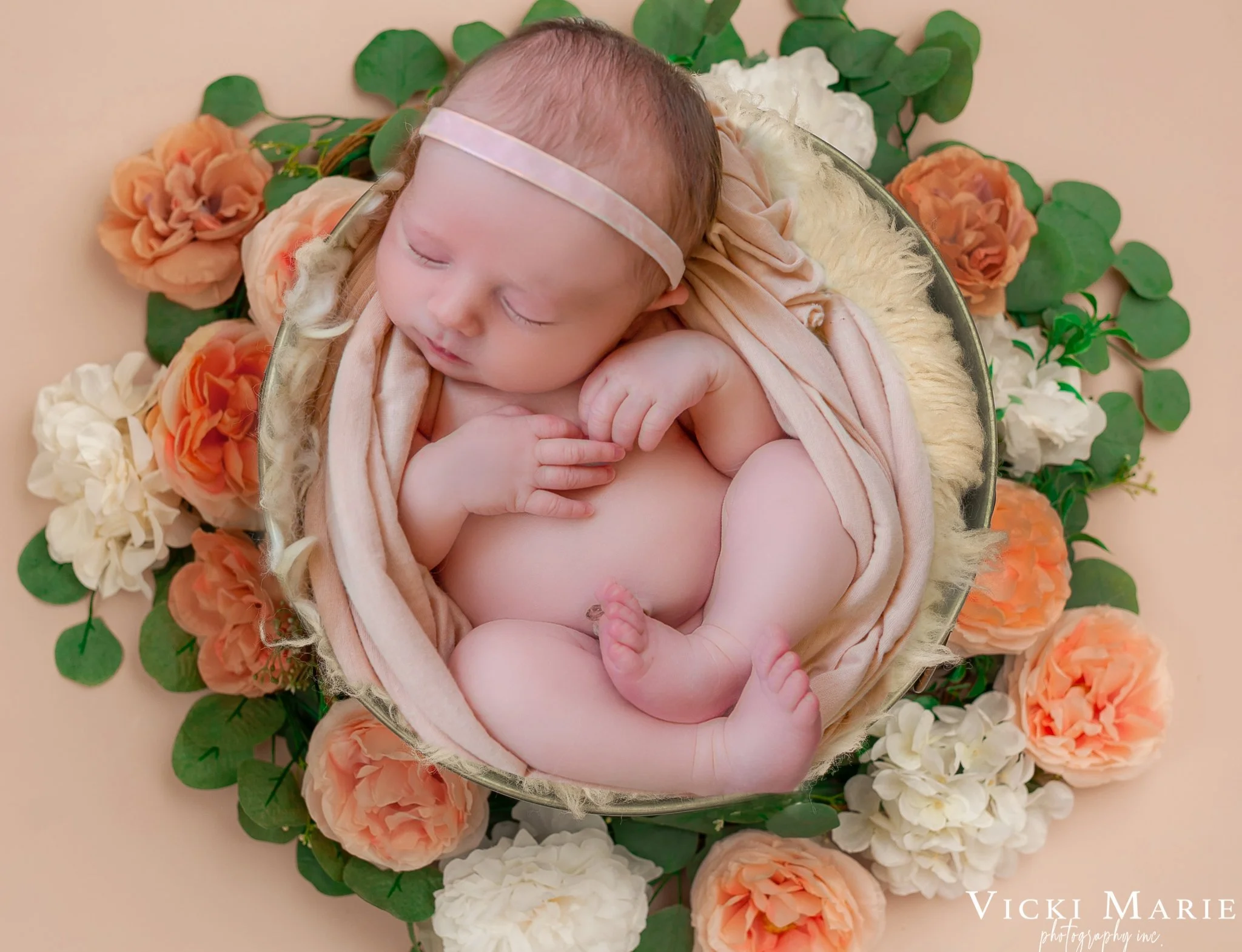 A sleeping newborn baby girl wrapped in a soft blush pink cloth, lying in a round glass bowl on a bed of peach, white, and green flowers.