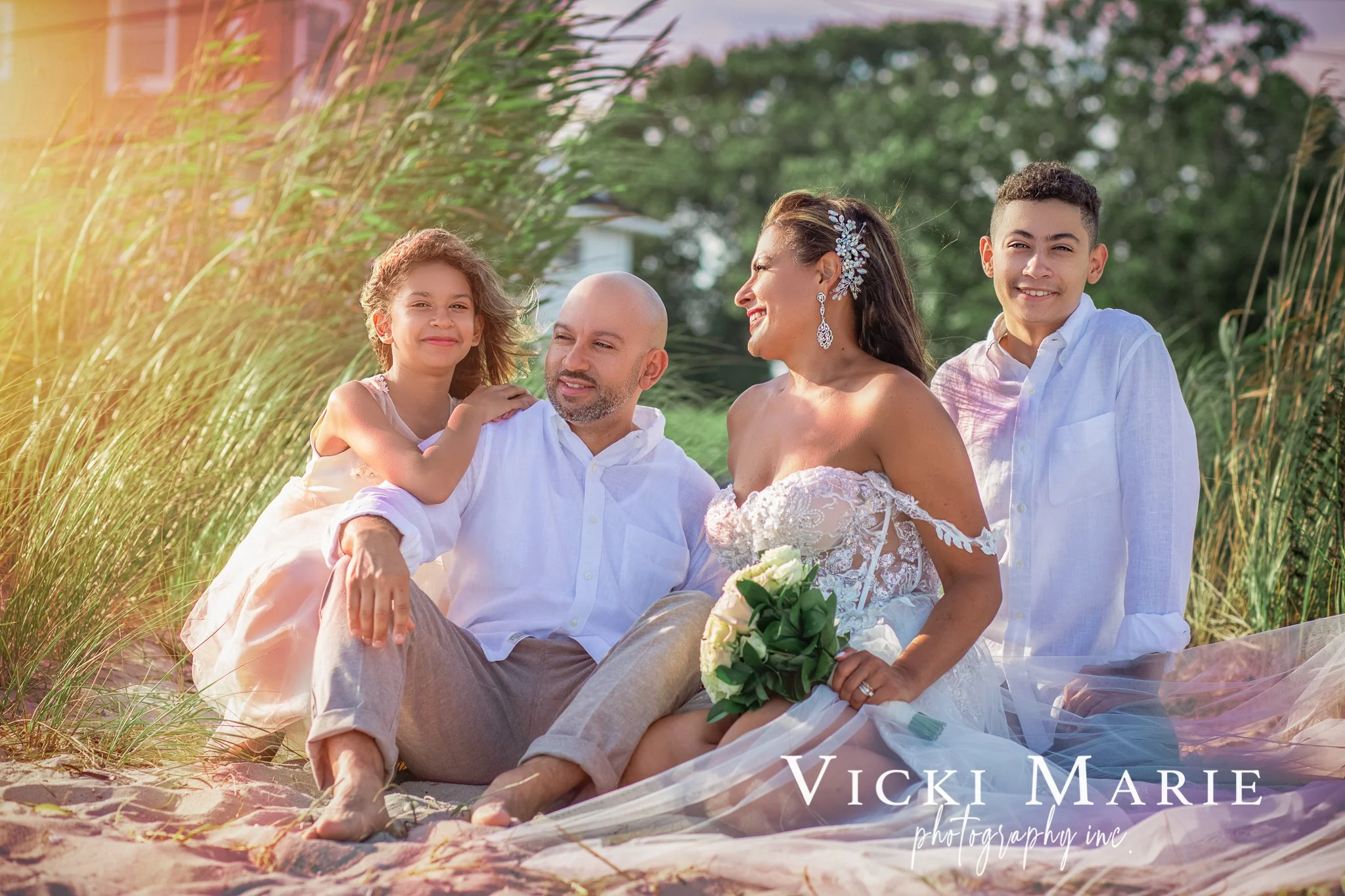 Wedding party sitting outdoors on a beach, including a bride in a wedding dress holding a bouquet, a groom, two children, and a woman, smiling and enjoying a sunny day.