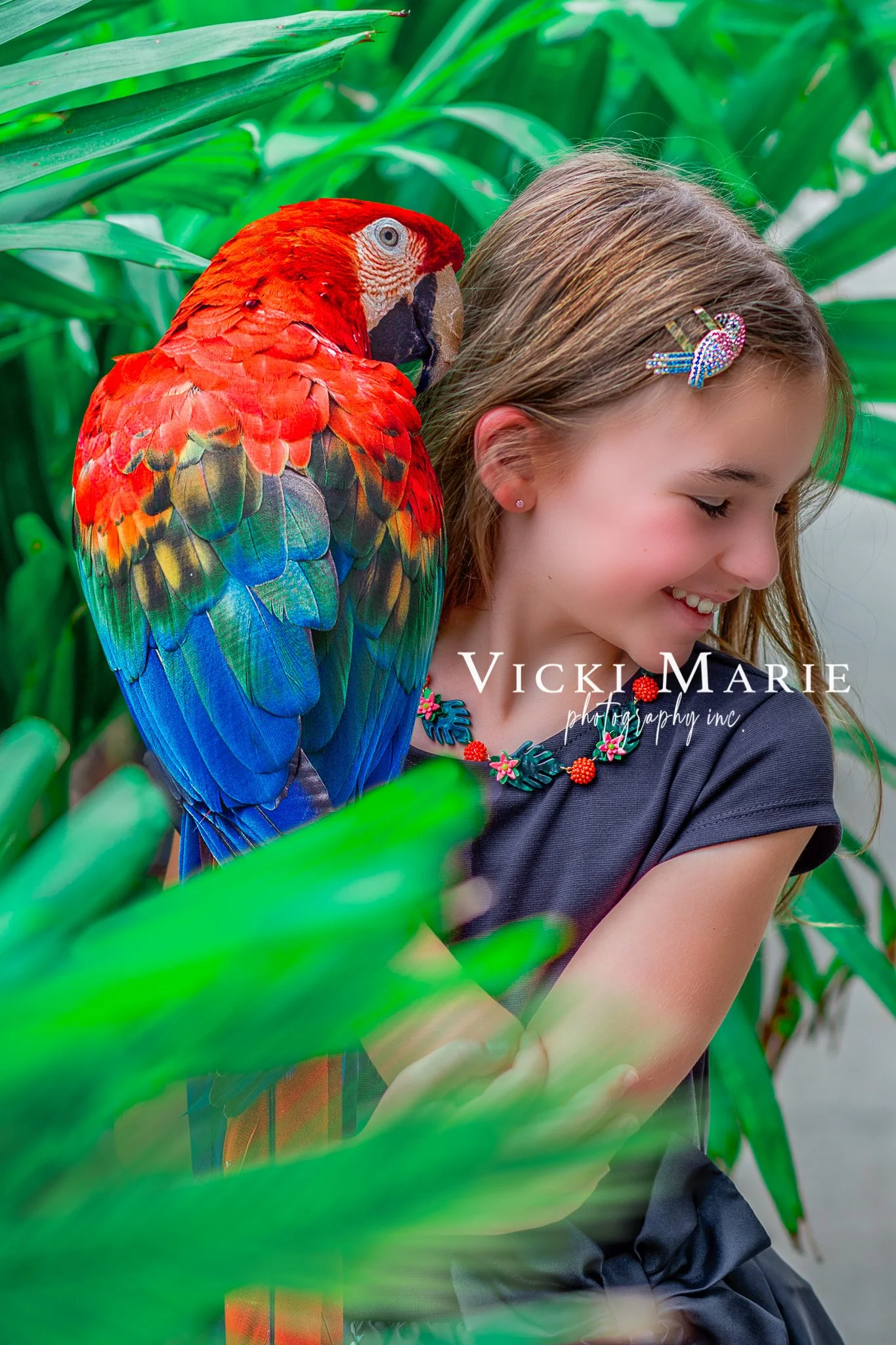 A young girl with long hair, wearing a dark dress and colorful jewelry, is smiling with her eyes closed as a large red, green, and blue parrot perches on her shoulder among green tropical plants.