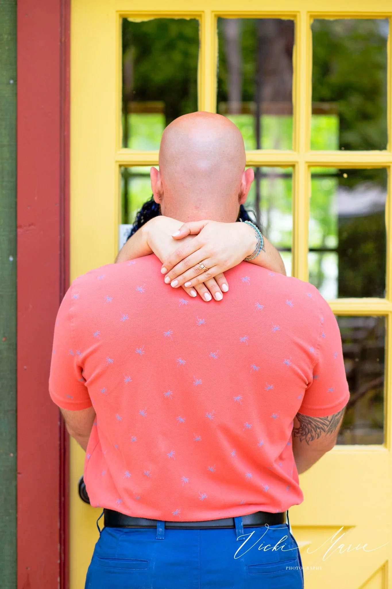 A couple hugging in front of a yellow door with a window. The person's head is bald and facing away, wearing an orange shirt with small patterns, while the woman has dark hair and is hugging from behind.