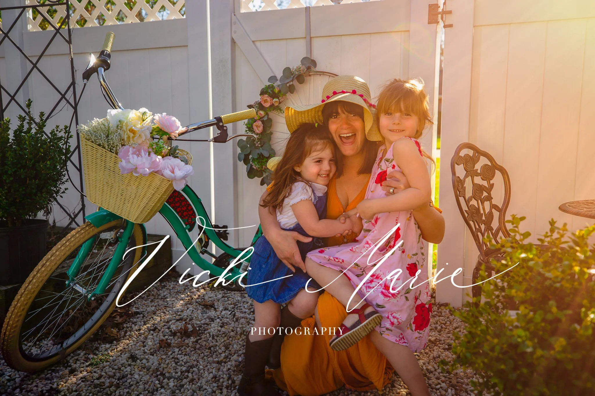A woman with two young girls smiling and hugging outdoors near a decorative white fence, a vintage bicycle with a flower basket, and a garden chair, during sunset.