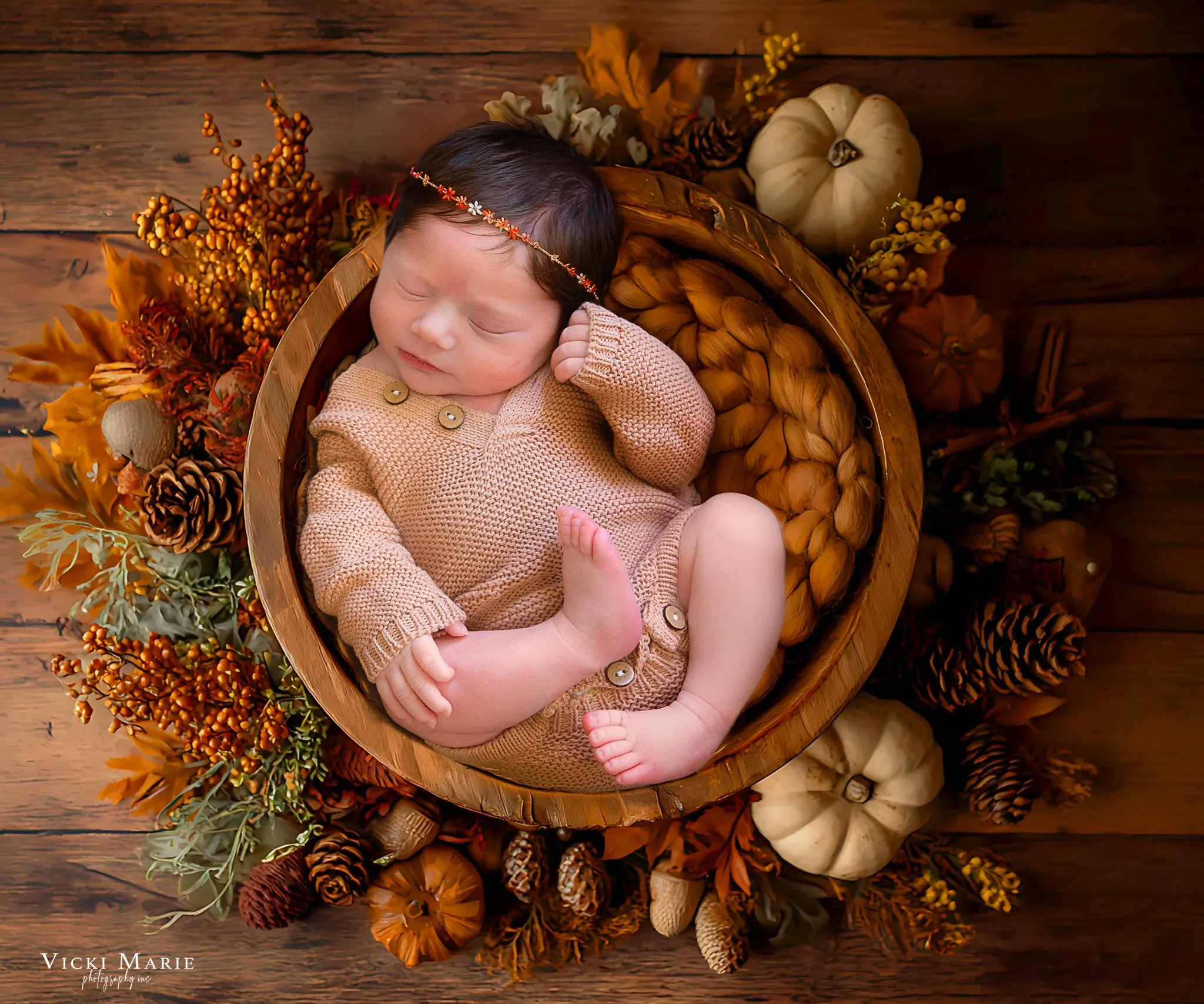 A sleeping baby with dark hair in a beige knitted outfit, lying in a round wooden basket, surrounded by fall-themed decorations like pumpkins, pinecones, and dried leaves on a wooden floor.