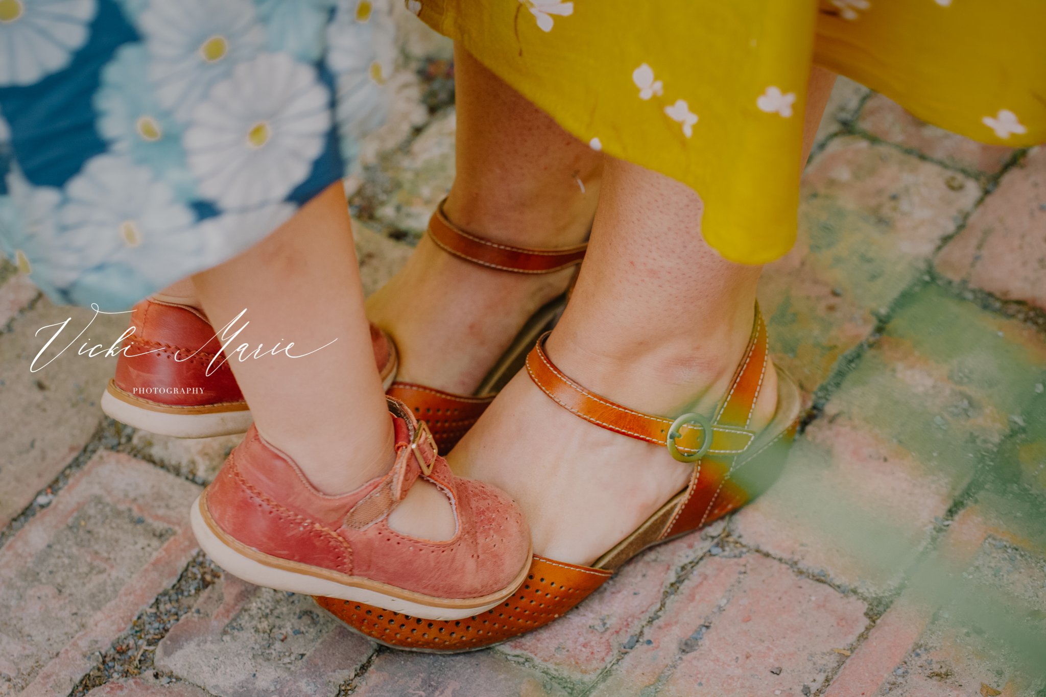 Close-up of two pairs of feet and shoes on brick pavement, one person wears pink shoes with cutouts and laces, the other wears orange-brown open-toe sandals with ankle straps, and part of a colorful floral dress is visible.