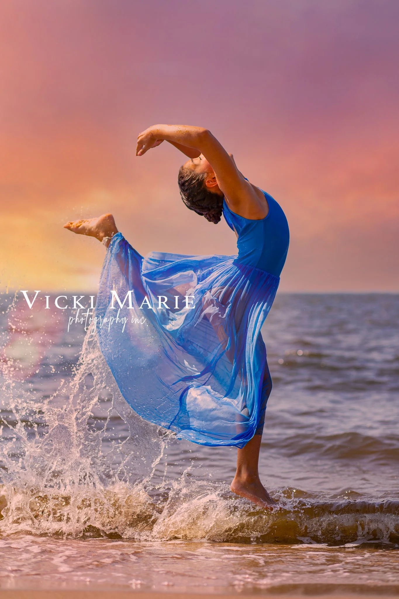 A woman in a blue dress dancing on the beach during sunset, splashing water as she lifts one leg.