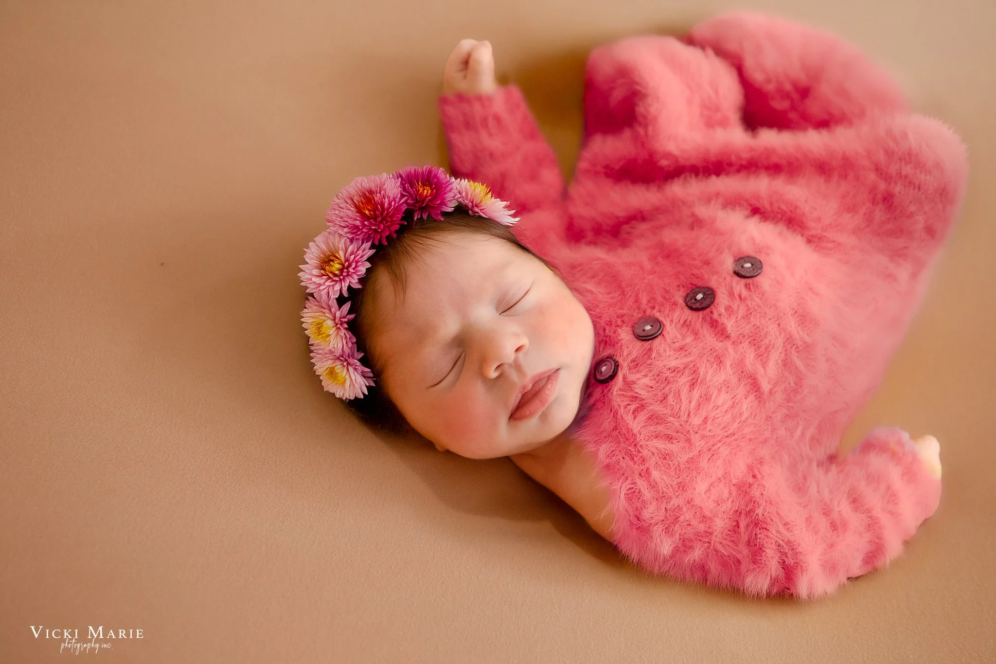A sleeping newborn baby girl wearing a pink fuzzy outfit and a floral headband, lying on a beige surface.