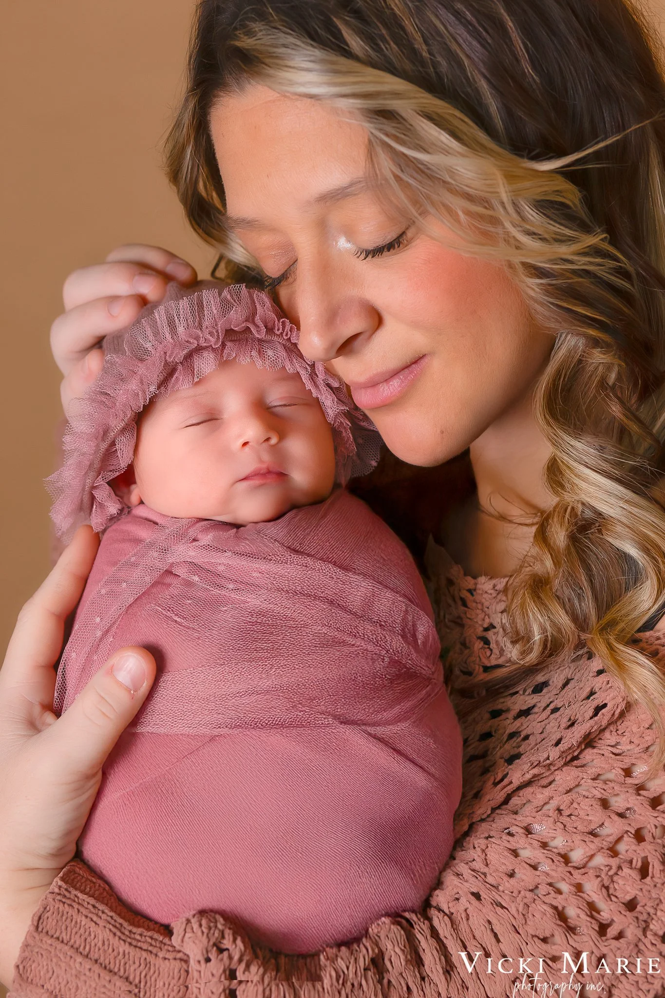 A woman with wavy, light brown hair and light skin gently holds and snuggles a sleeping baby wrapped in pink fabric and wearing a pink bonnet. The woman has her eyes closed and a tender expression.