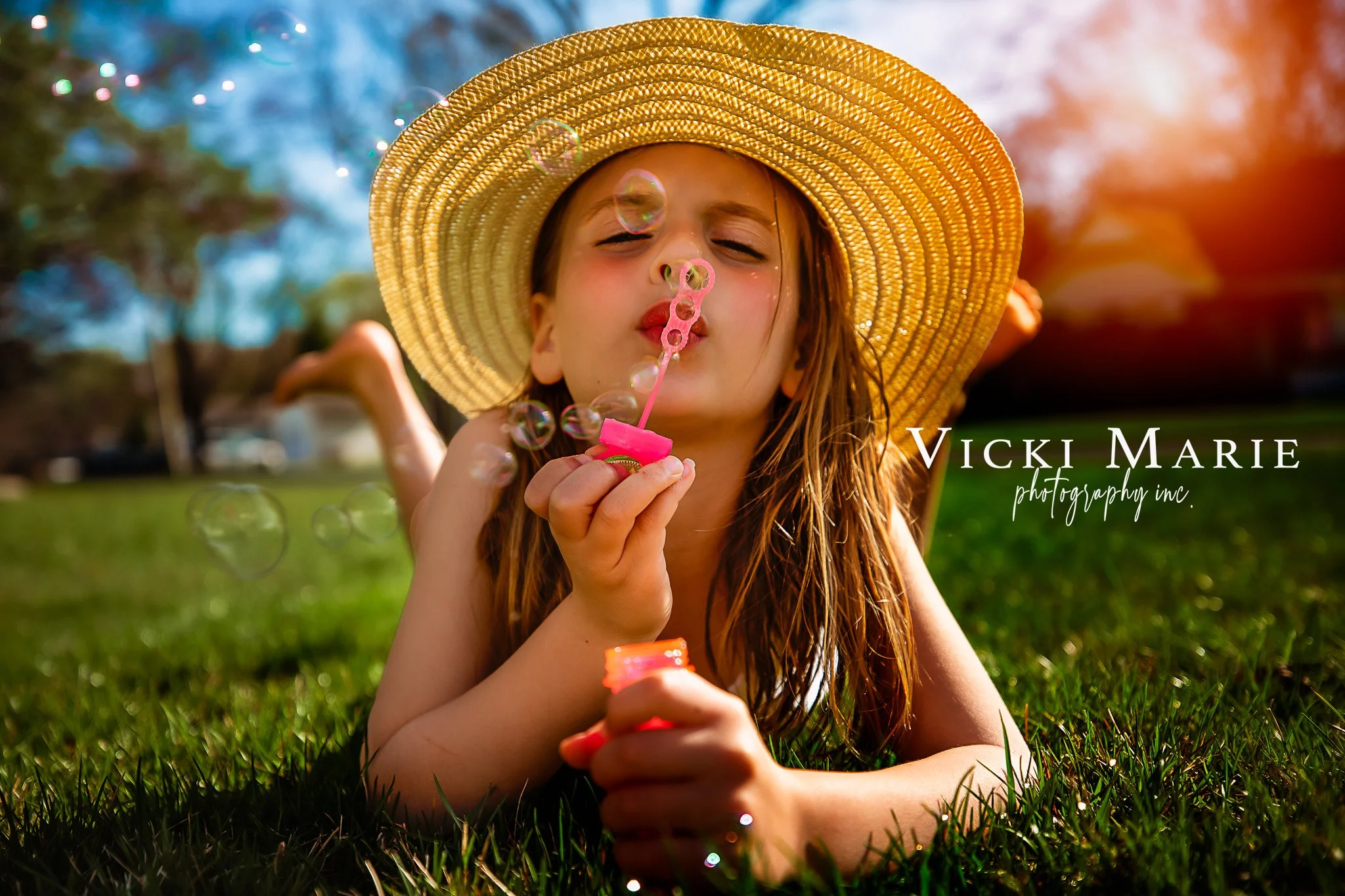 Young girl lying on grass, blowing bubbles, wearing a wide-brimmed straw hat, with sunlight and blue sky in the background.