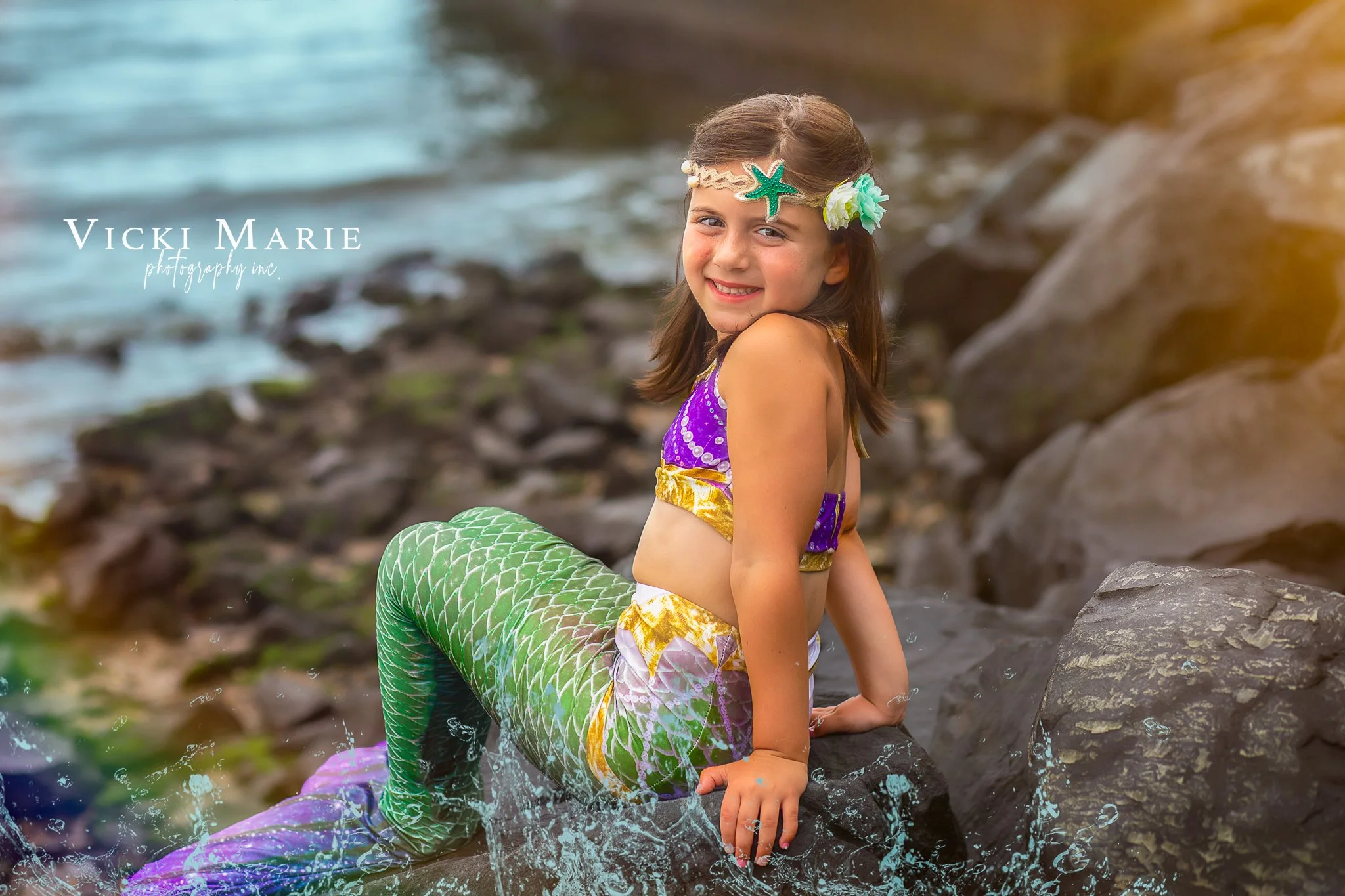 A young girl dressed as a mermaid sitting on rocks by the water, smiling at the camera. She is wearing a purple and yellow mermaid costume with a green fish tail, a shell headband, and flower accessories in her hair.