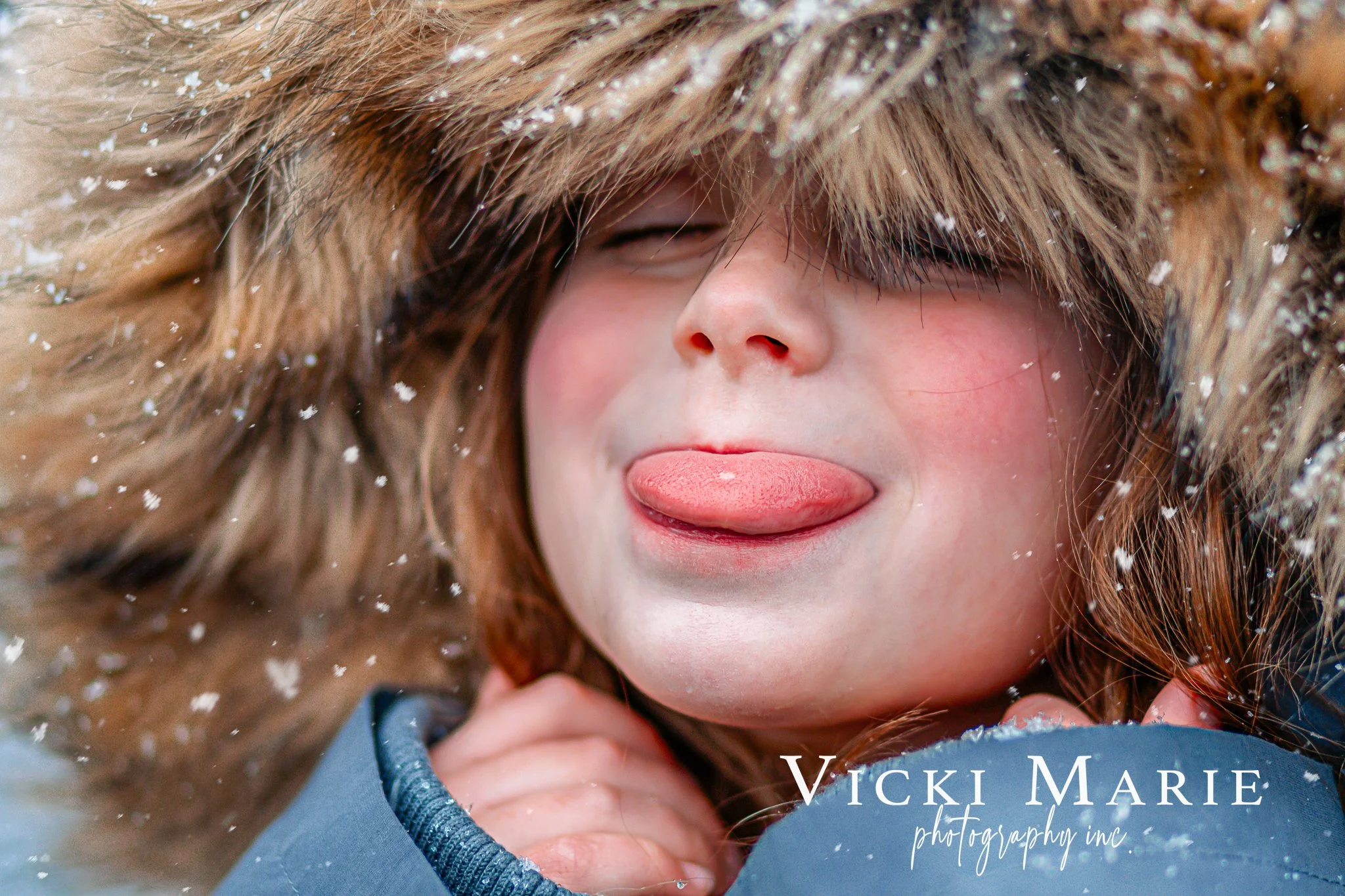 A young girl with light skin and long, wavy brown hair, wearing a winter jacket with a furry hood, sticking her tongue out and squinting her eyes, surrounded by snowflakes.