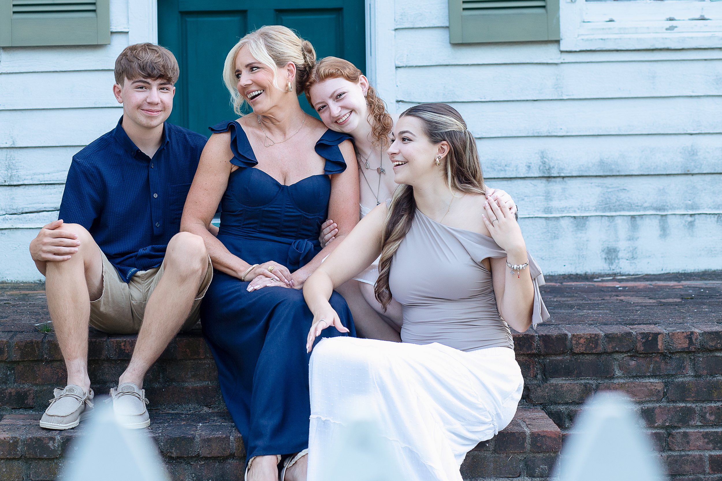 A woman sitting on brick steps with three young women, all smiling and laughing, with a white wooden house in the background.
