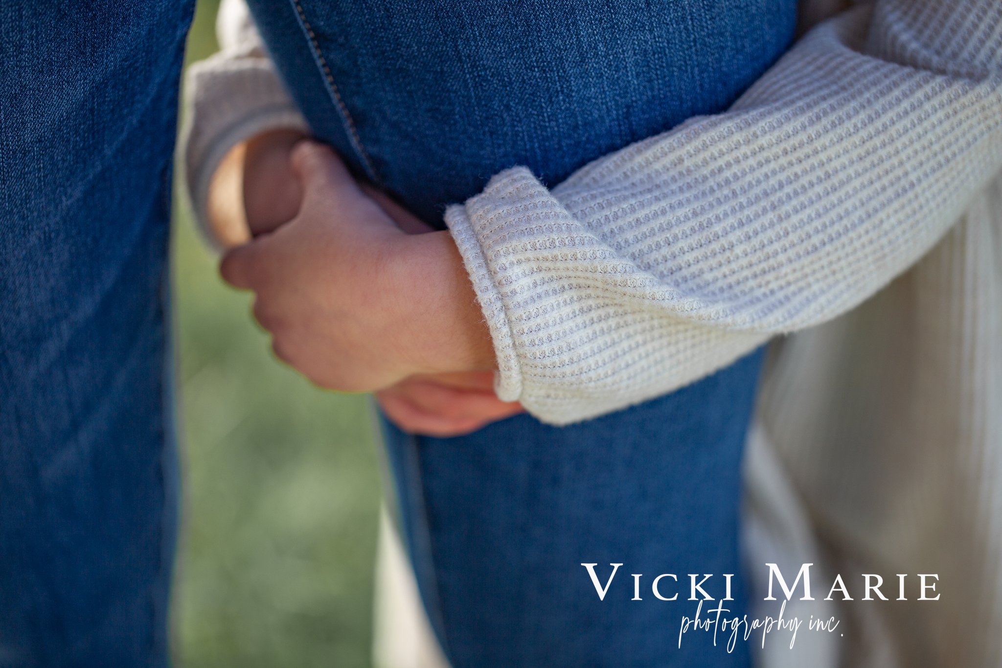 A person wearing a gray long-sleeve shirt holding a child, whose arm is wrapped around the person's waist. The person is wearing blue jeans, and the child is wearing a shirt and beige pants. The background is blurred.