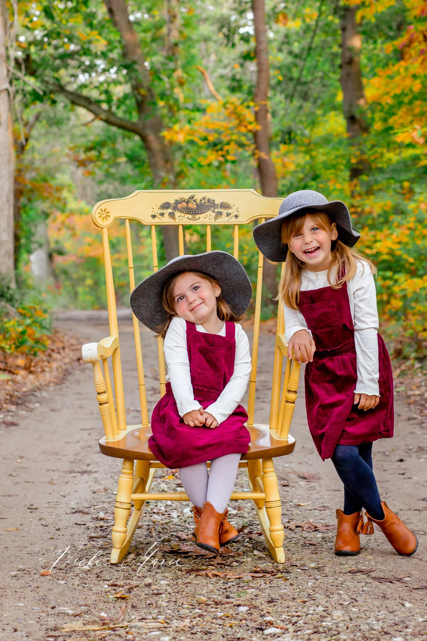 Two young girls wearing gray wide-brim hats and matching maroon dresses with white long sleeve shirts underneath, posing outdoors on a dirt path surrounded by trees with autumn foliage. One girl is sitting on a yellow wooden rocking chair, and the ot